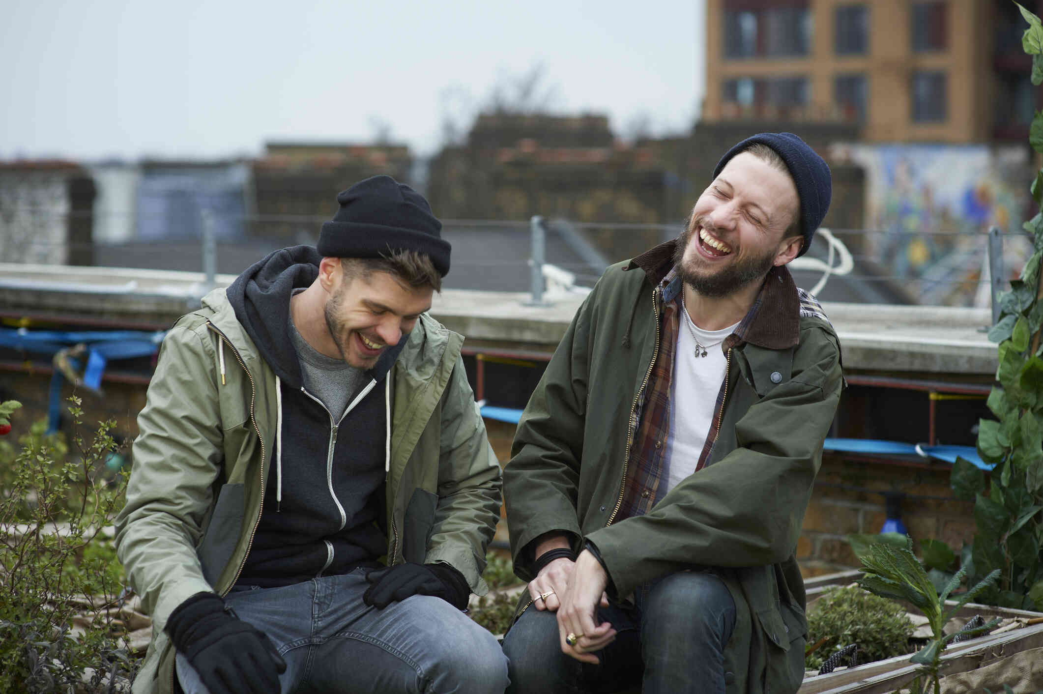 Two men wearing beanies sit next to each other on a patio while chattnig and smiling.