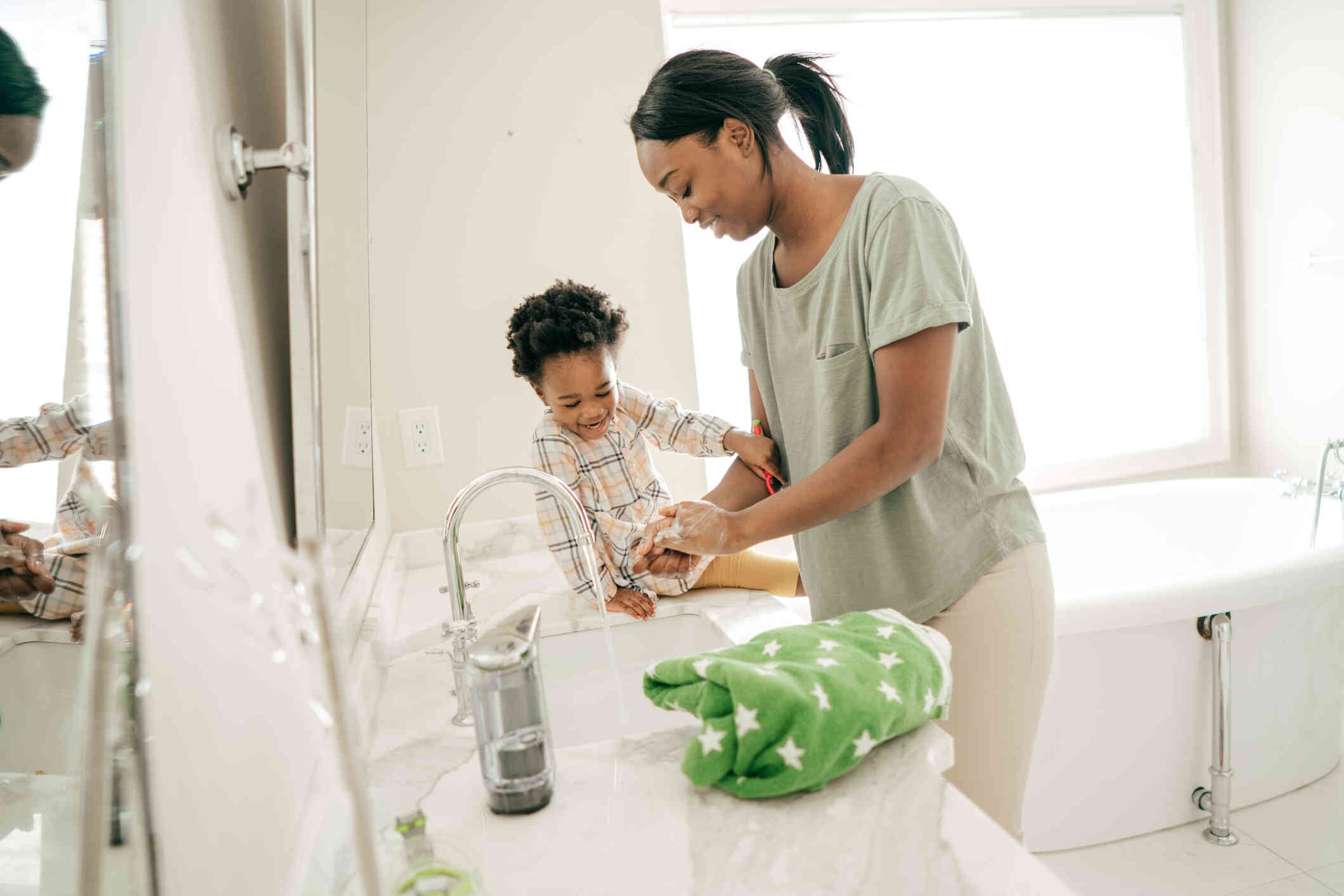 A mother stands in the bathroom washing her hands as her toddler son sits on the coucnter next to her laughing.