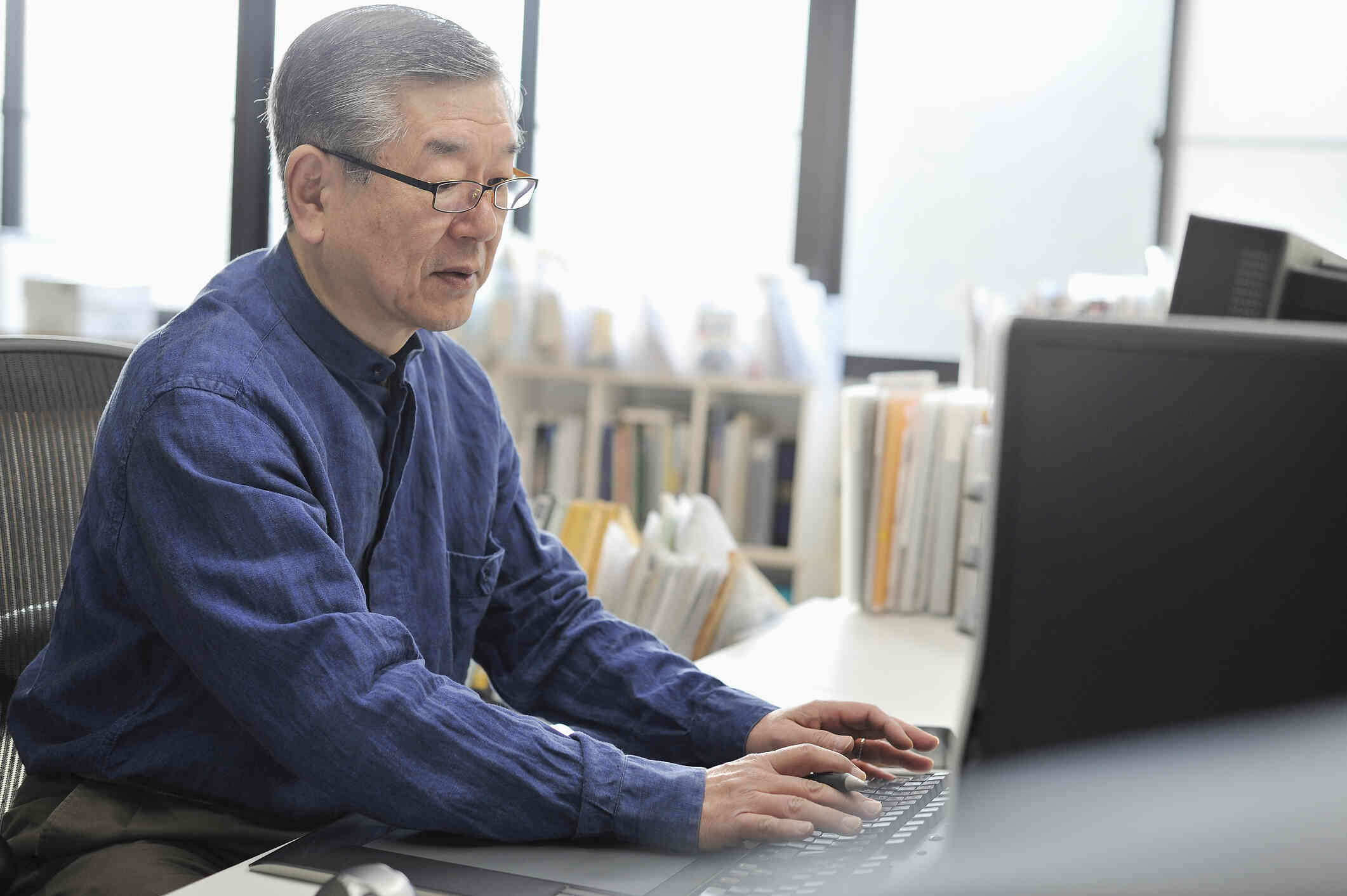 A mature man in a blue shirt sits at his desk at work and looks at the computer infornt of him with serious expression.