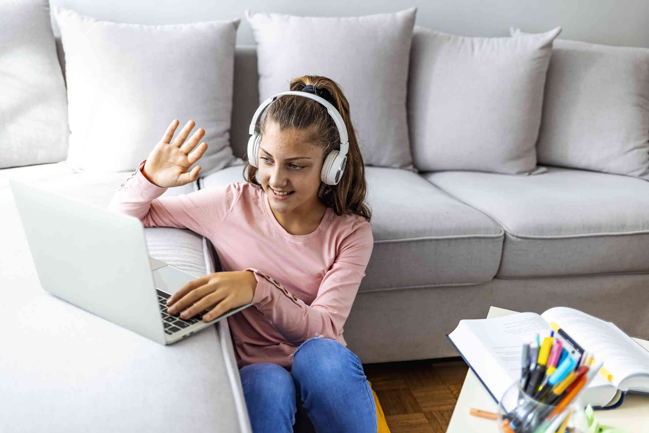 A teen girl in a pink shirt sits on the floor next to the couch while wearing white headphones as she waves at the laptop screen on the couch infront of her with her study books on the coffee table.