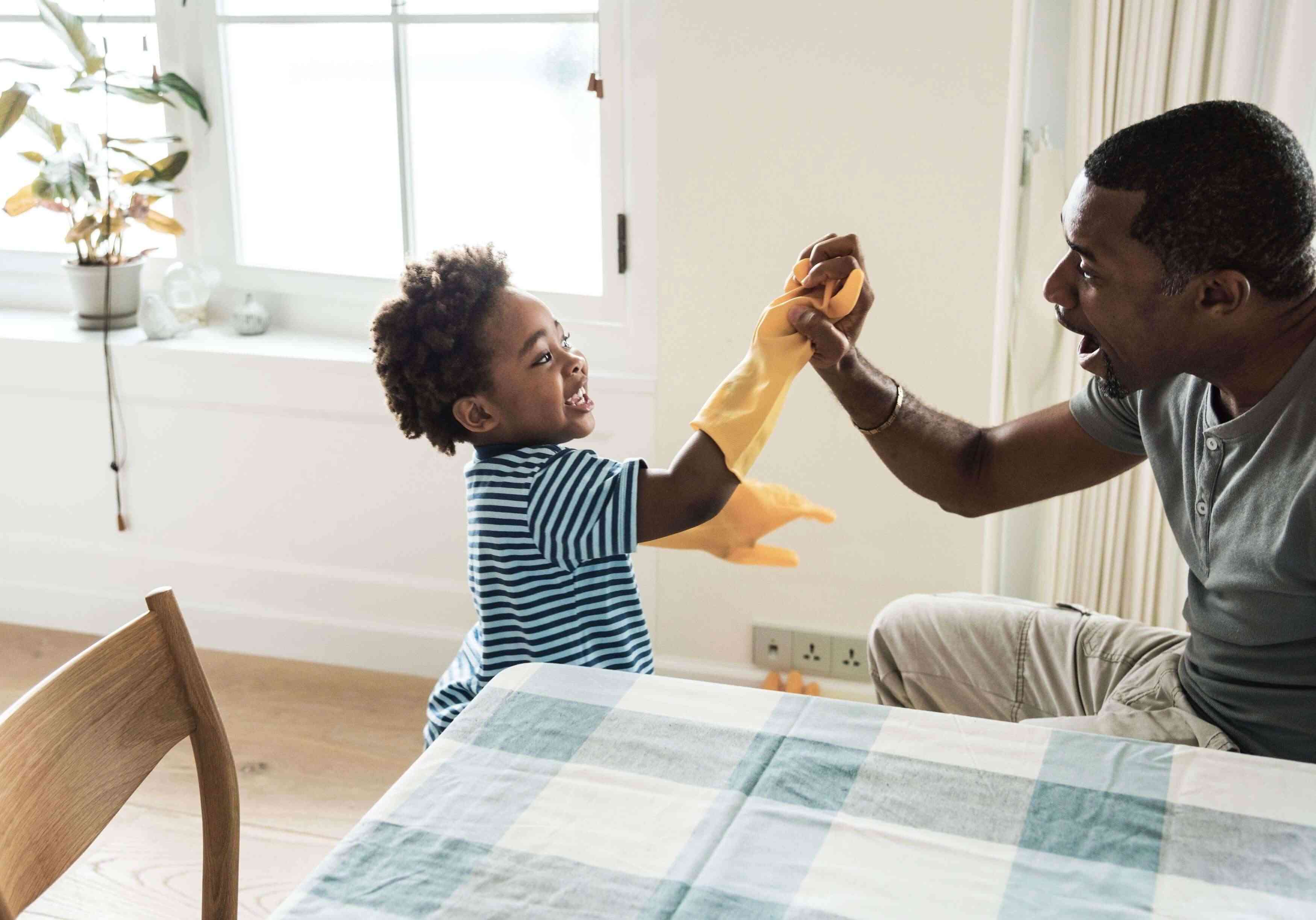 A dad high fives his young son who is wearing a large cleaning glove as he teaches his child how to do chores.