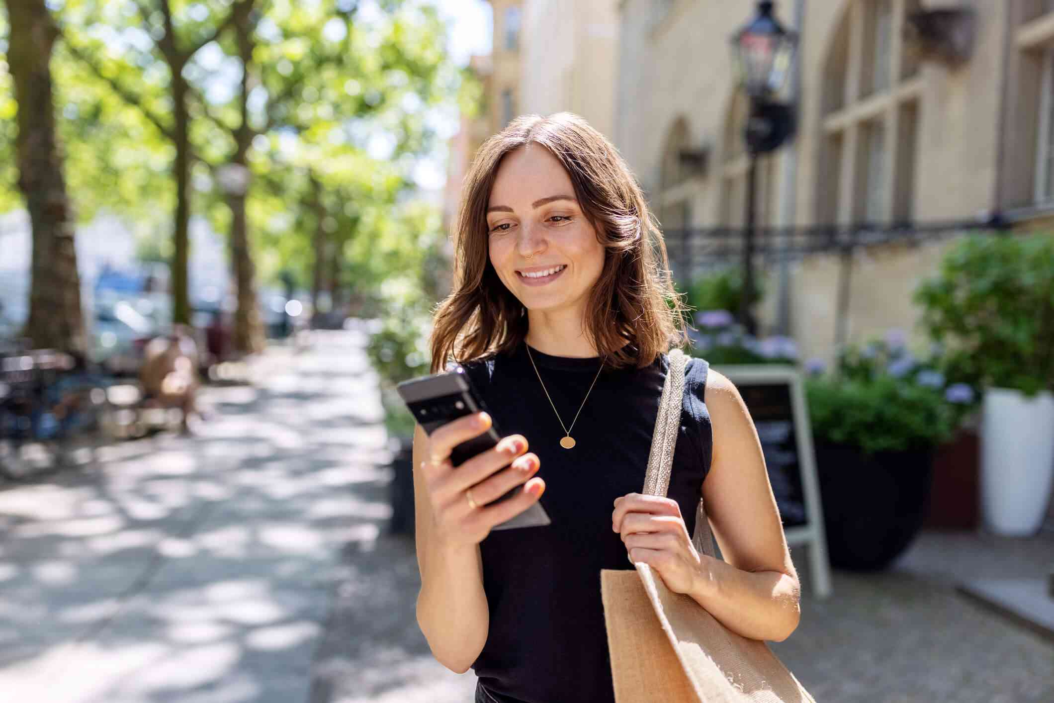 A woman in a black shirt stands otuside and smiles down at her phone in her hand on a sunny day.
