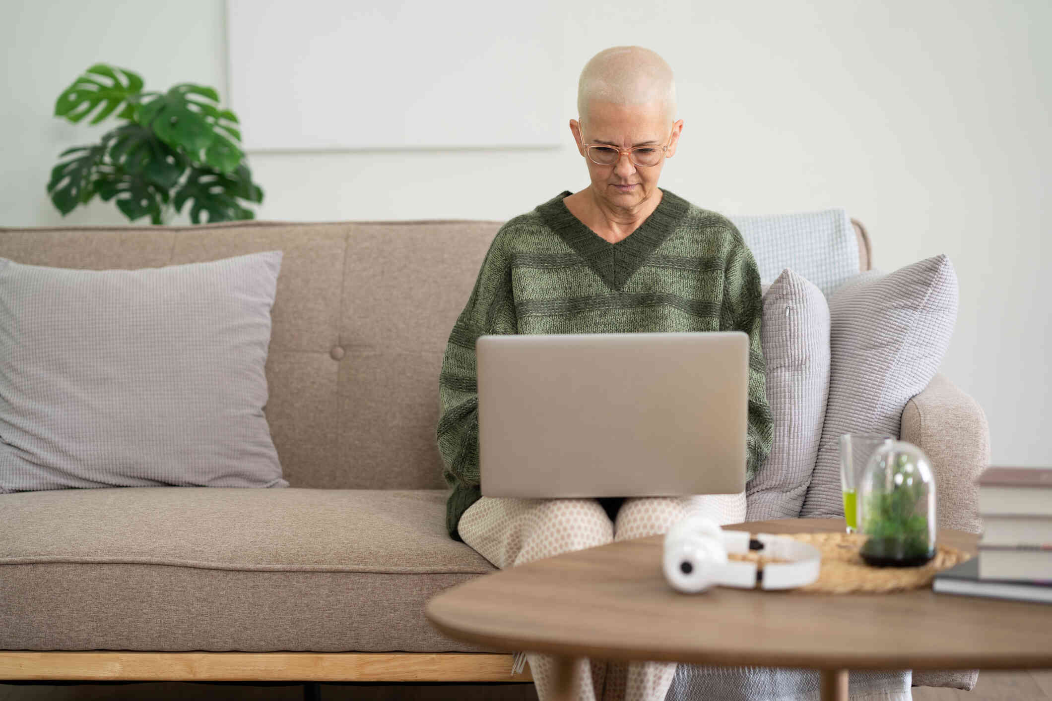 A mature woman in a green sweater sits on a couch in her living room and looks down at the laptop open in her lap with a serious expression.