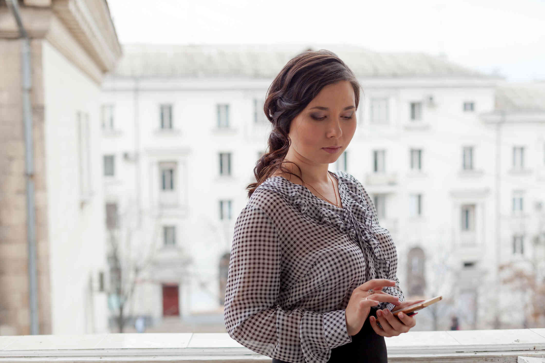 A woman in a checkerd shirt stands infront of a window and looks down at the cellphone in her hand.