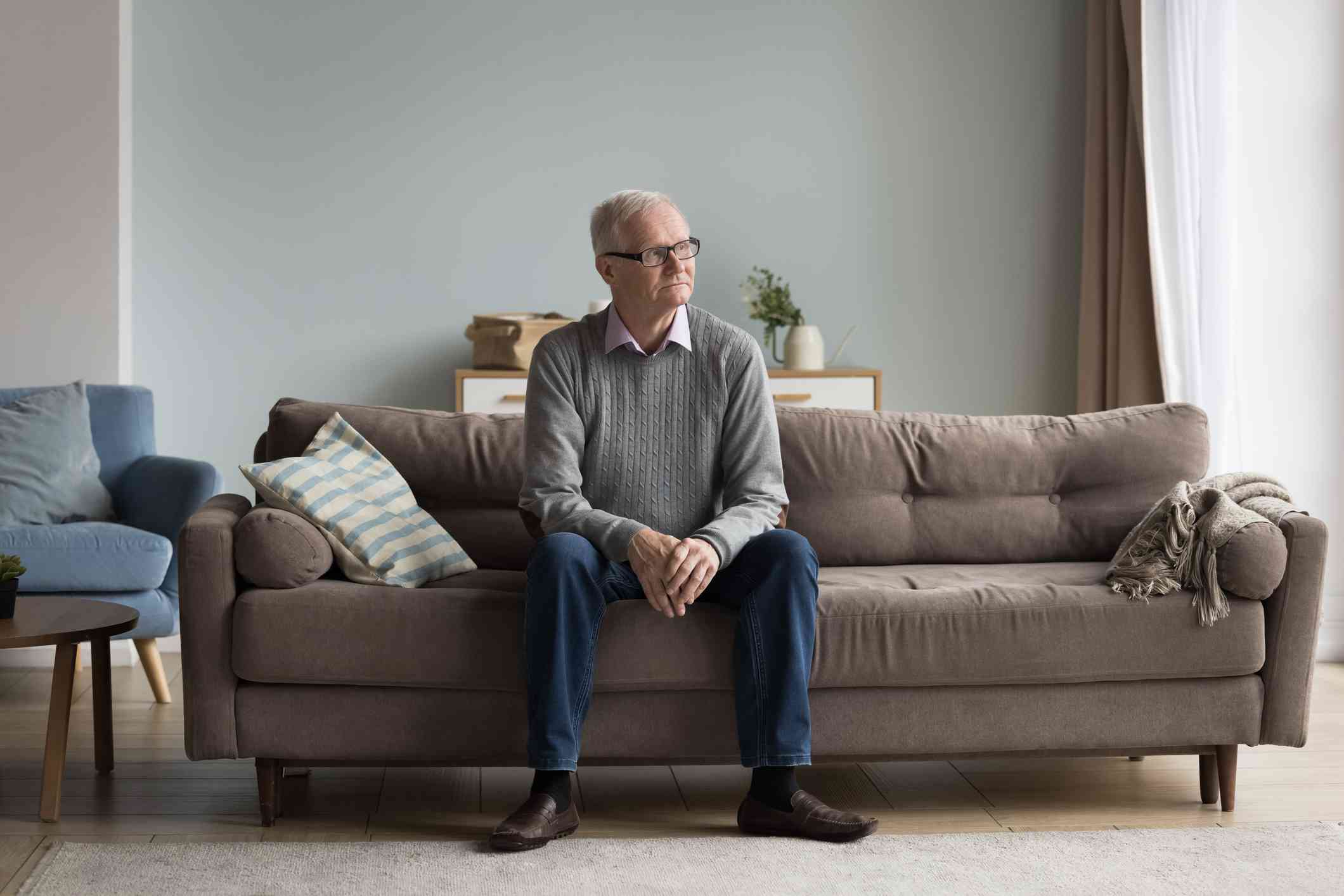 An elderly man in a grey sweater sits on the couch in his living room and gazes off with a sad expression.