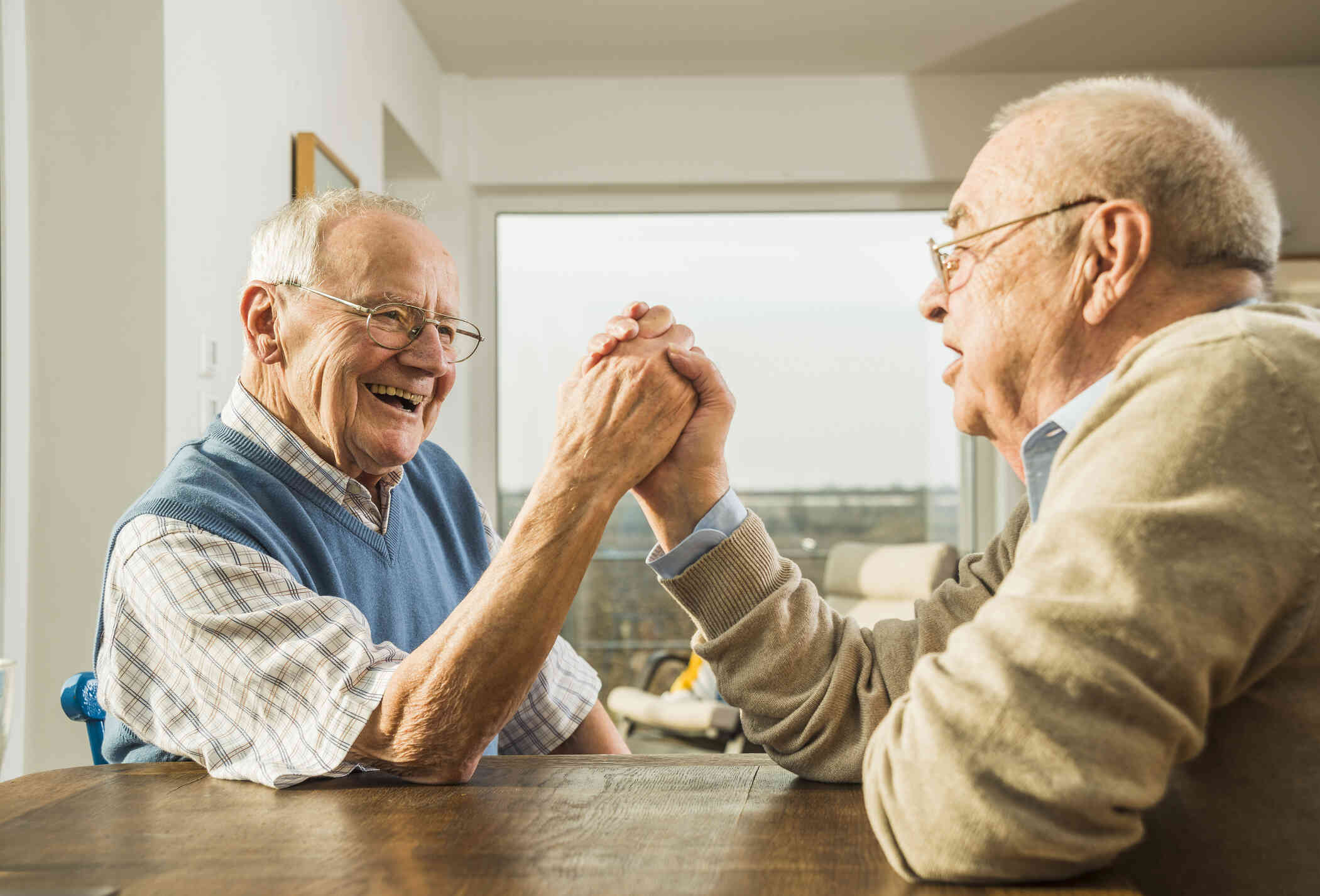 Two elderly man sit across from each other at a wooden table and clasp each others hand while smiling at one another.