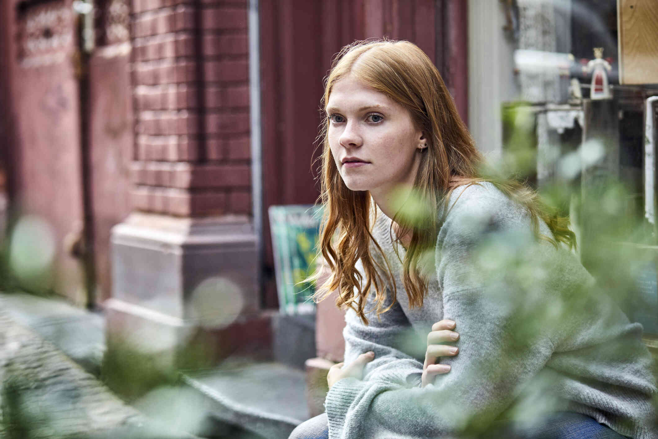 A woman in a grey sweater sits hunched over on a step outside of a brick building while gazing off.