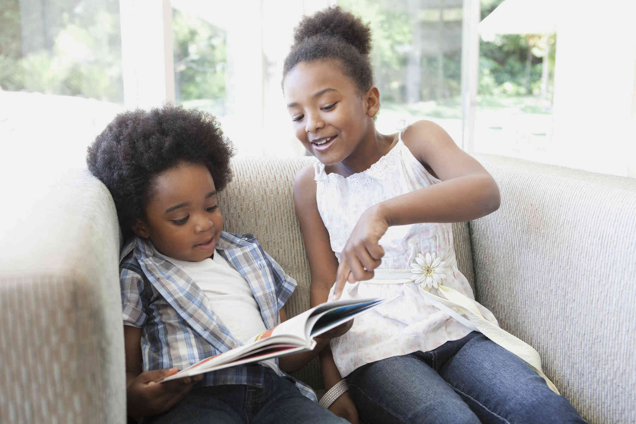 A young girl sits on a couch and point to a spot on a book in her toddler brothers lap.