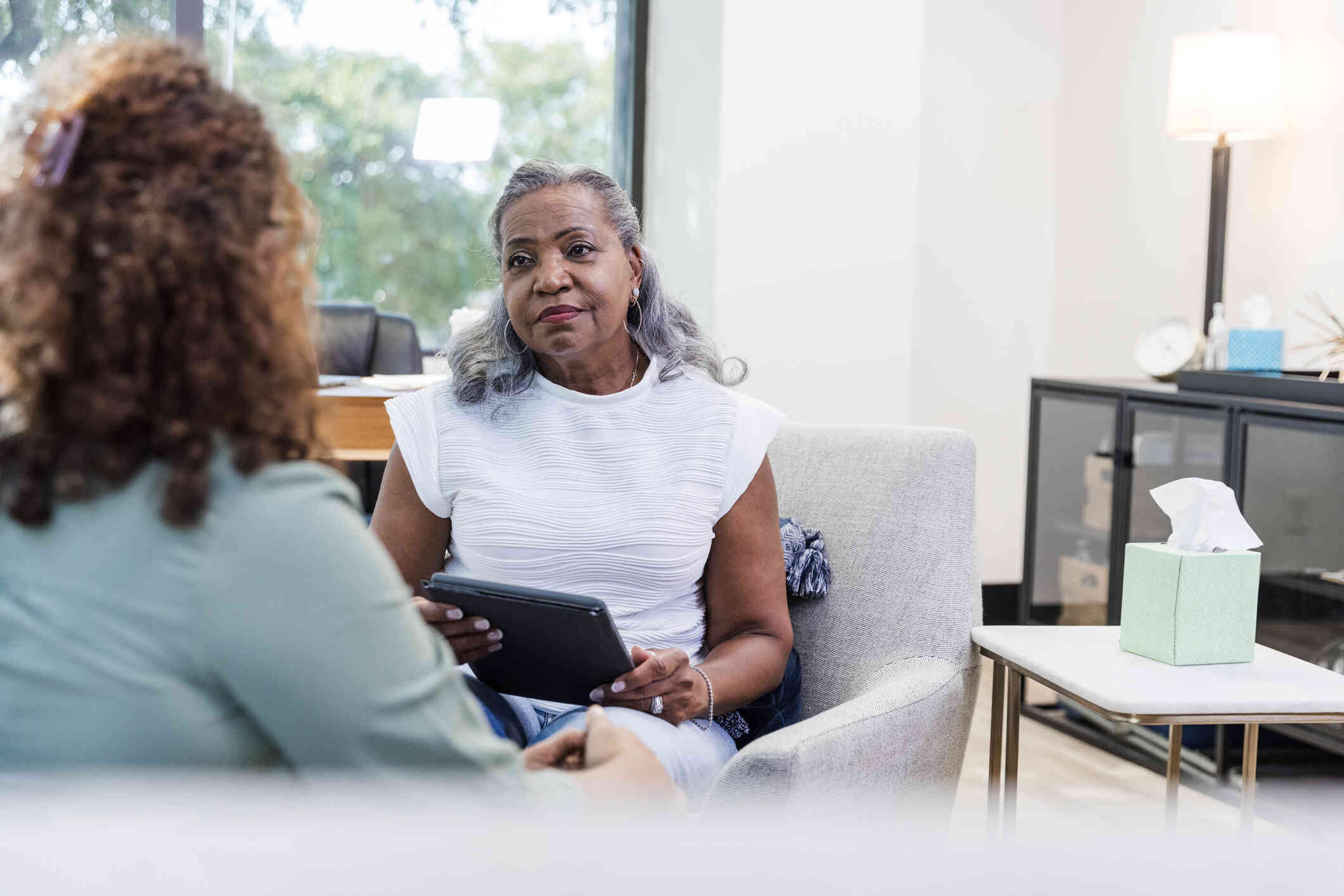 A female therapist with a clipboard listens intently to the female patient sitting across form her during a therapy session.