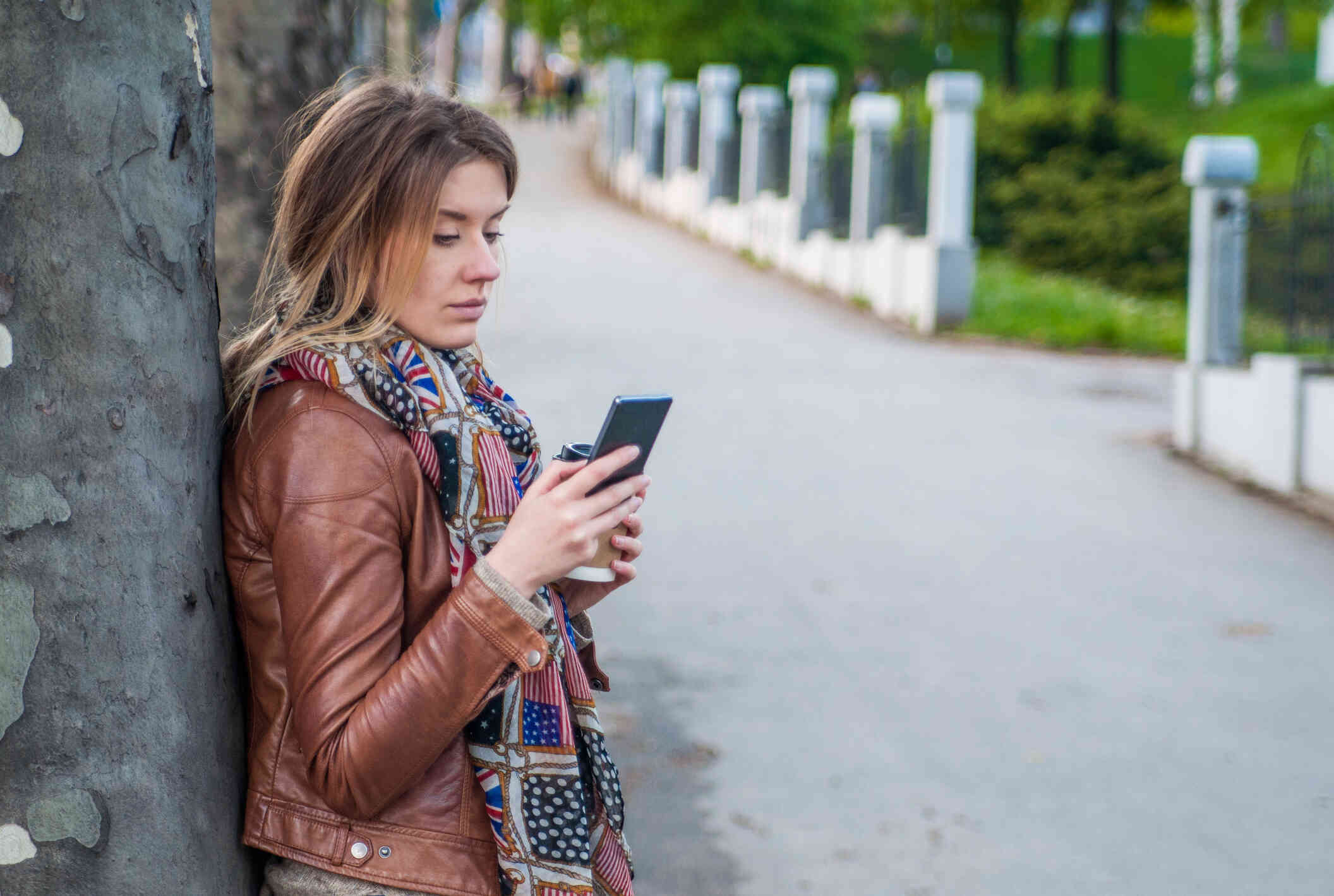 A woman in a brown leather jacket stands outside with a to go cup of coffee and looks at the cellphone in her hand.