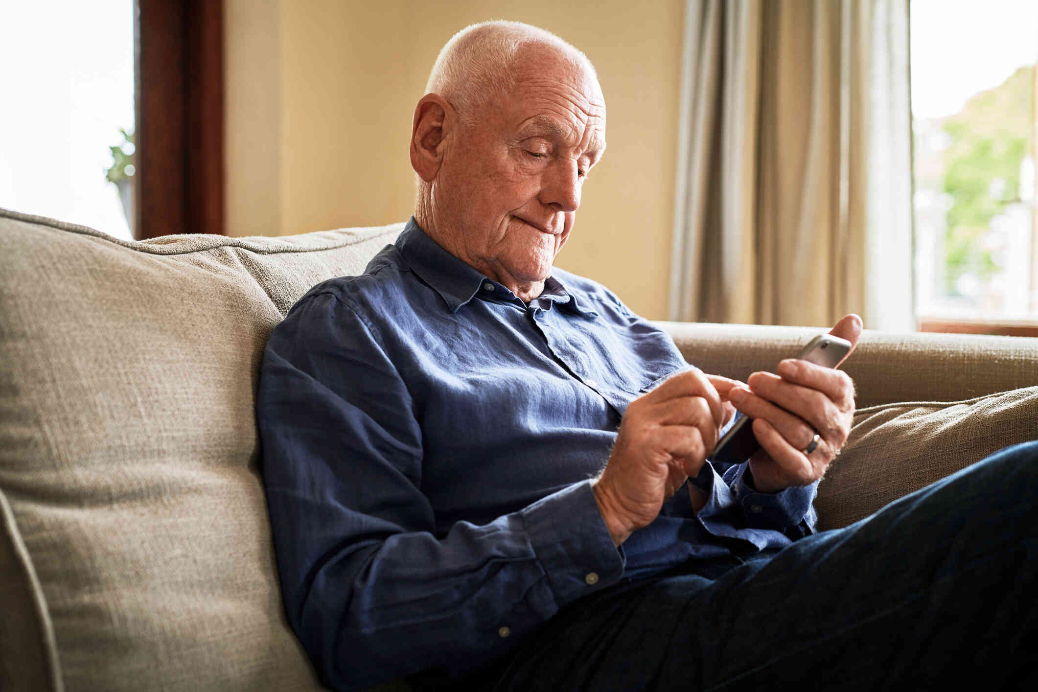 An elderly man in a blue button down shirt sits on the couch in his home and uses the cellphone in his hand.