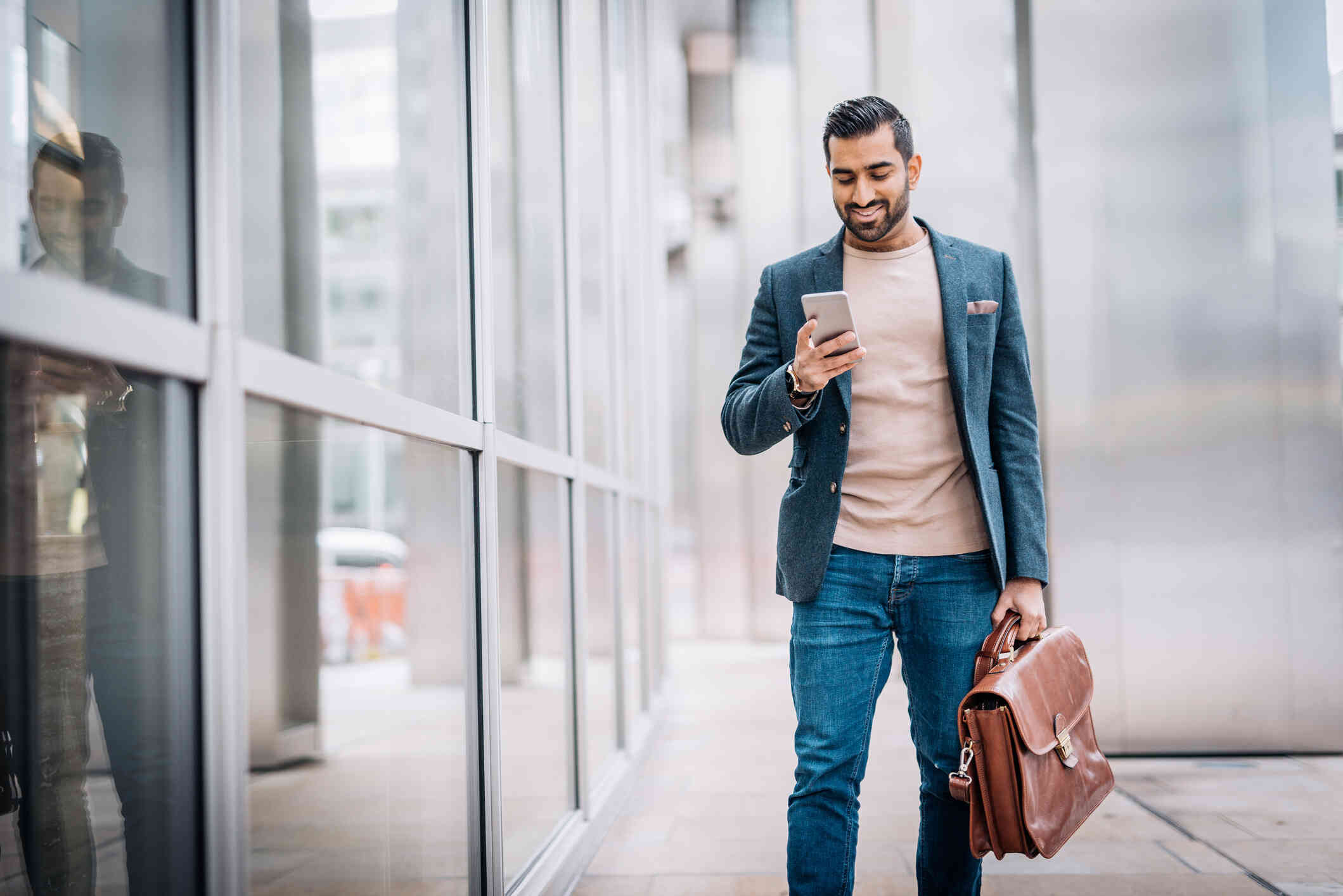 A man with a brown briefcase walks outside near a large building and smiles down at the phone in his hand.