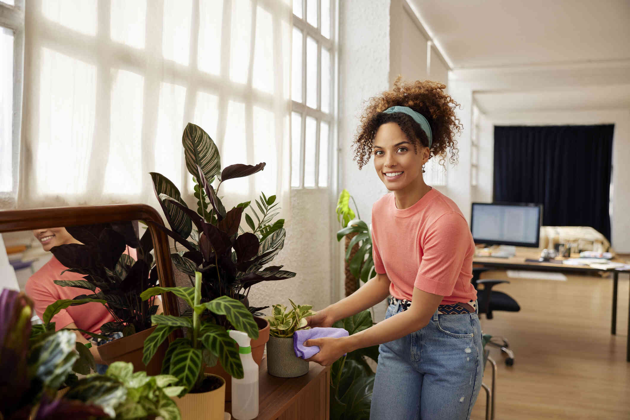 A woman in a pink shirt tends to her collection of indoor plants while smiling at the camera.