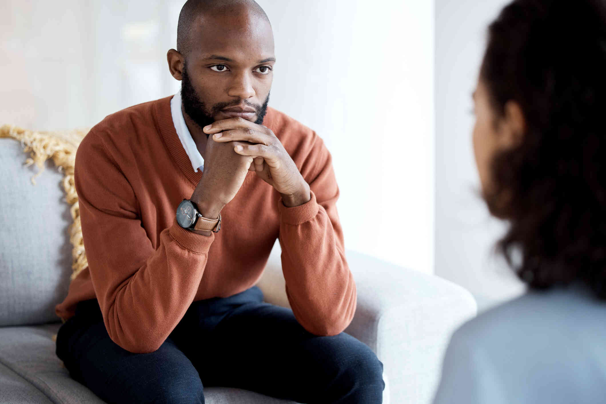 A man in a green sweater leans forward while sitting on the couch while pressing his hands together as he listens to the therapist sitting across from him.