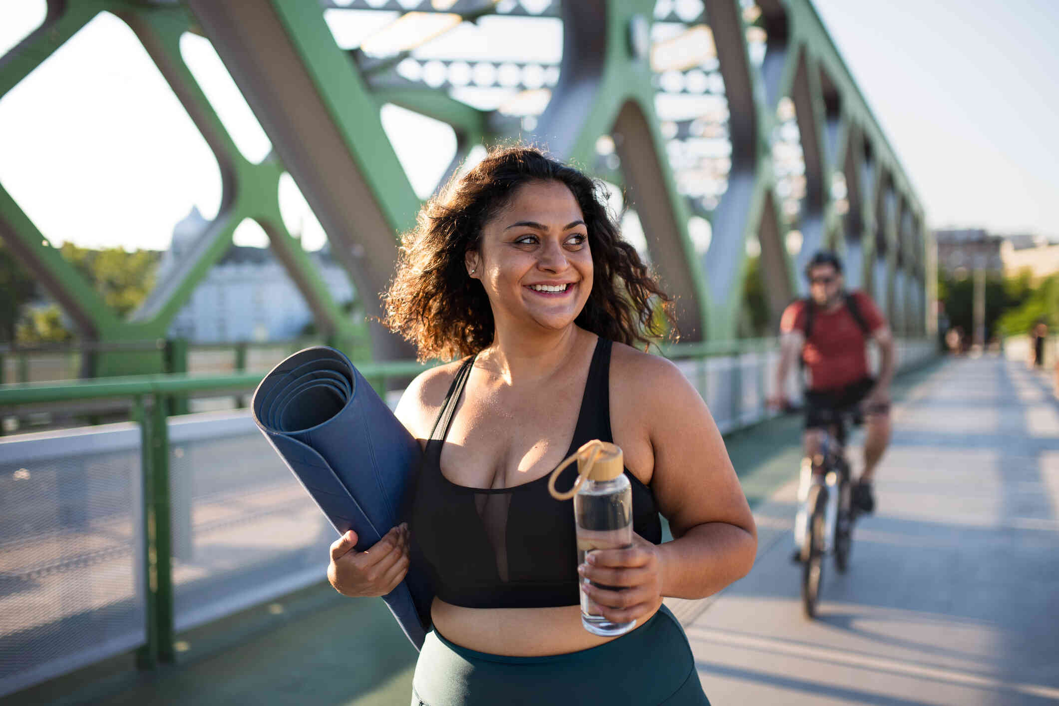 A woman in workout clothes walks along a bridge with a smile while holding a yoga mat and a water bottle.