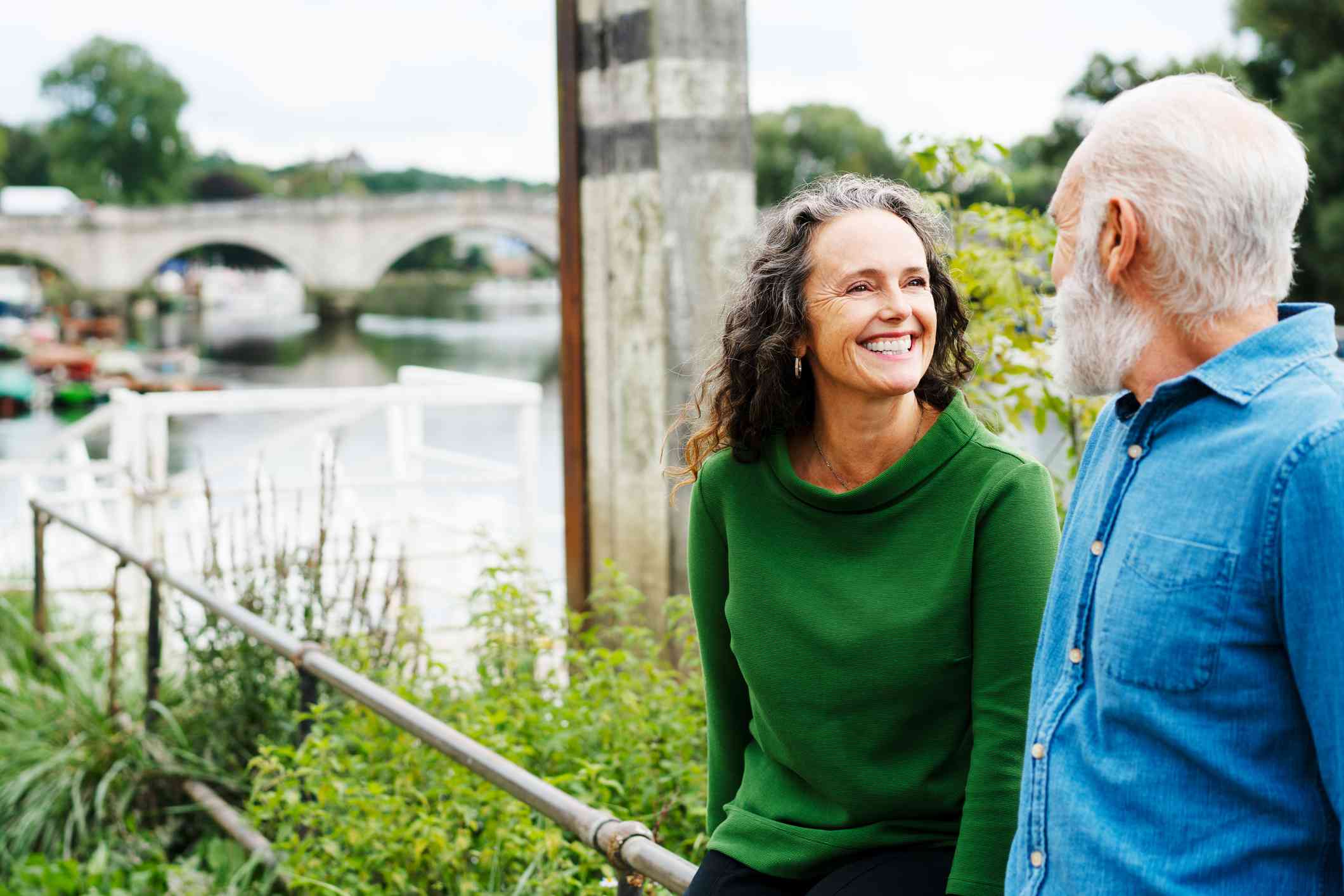 A mature male and female couple lean against a raiiling  while outside on a sunny day and smile at one another.