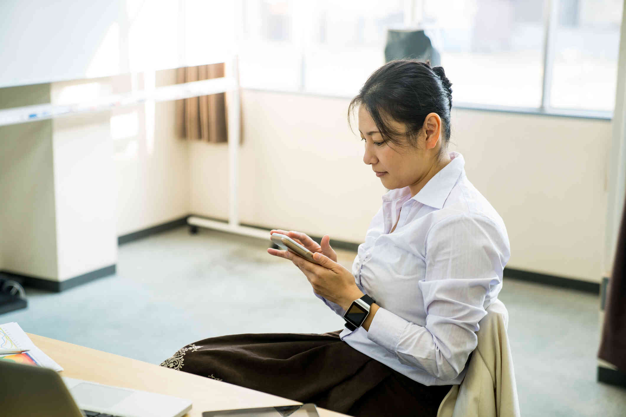 A woman in a white shirt sits in a chair and looks down at the phone in her hand with a serious expression.