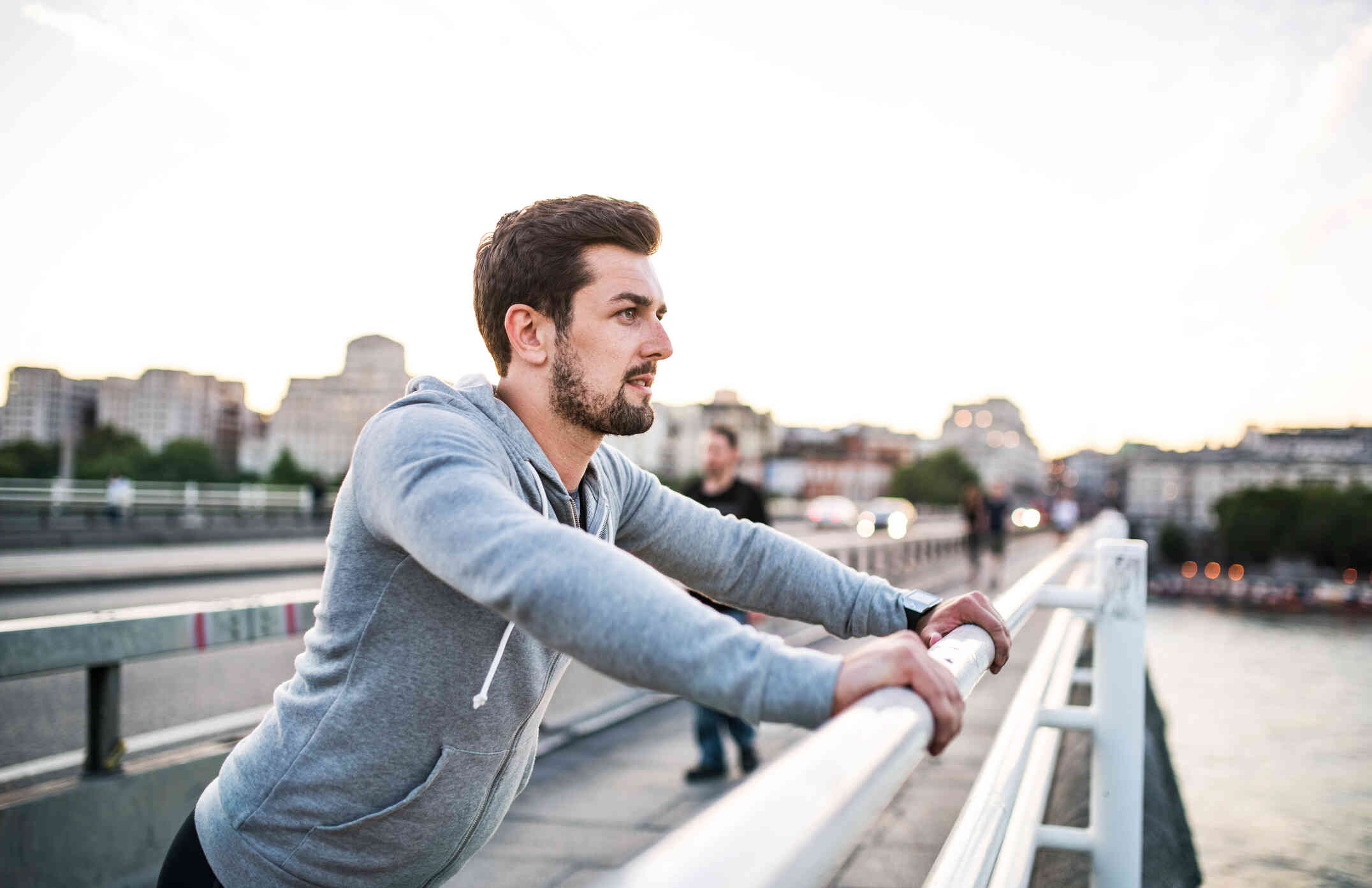 A man with a beard wears a gray hoodie as he looks out with a serious expression and leans against a metal railing outside.