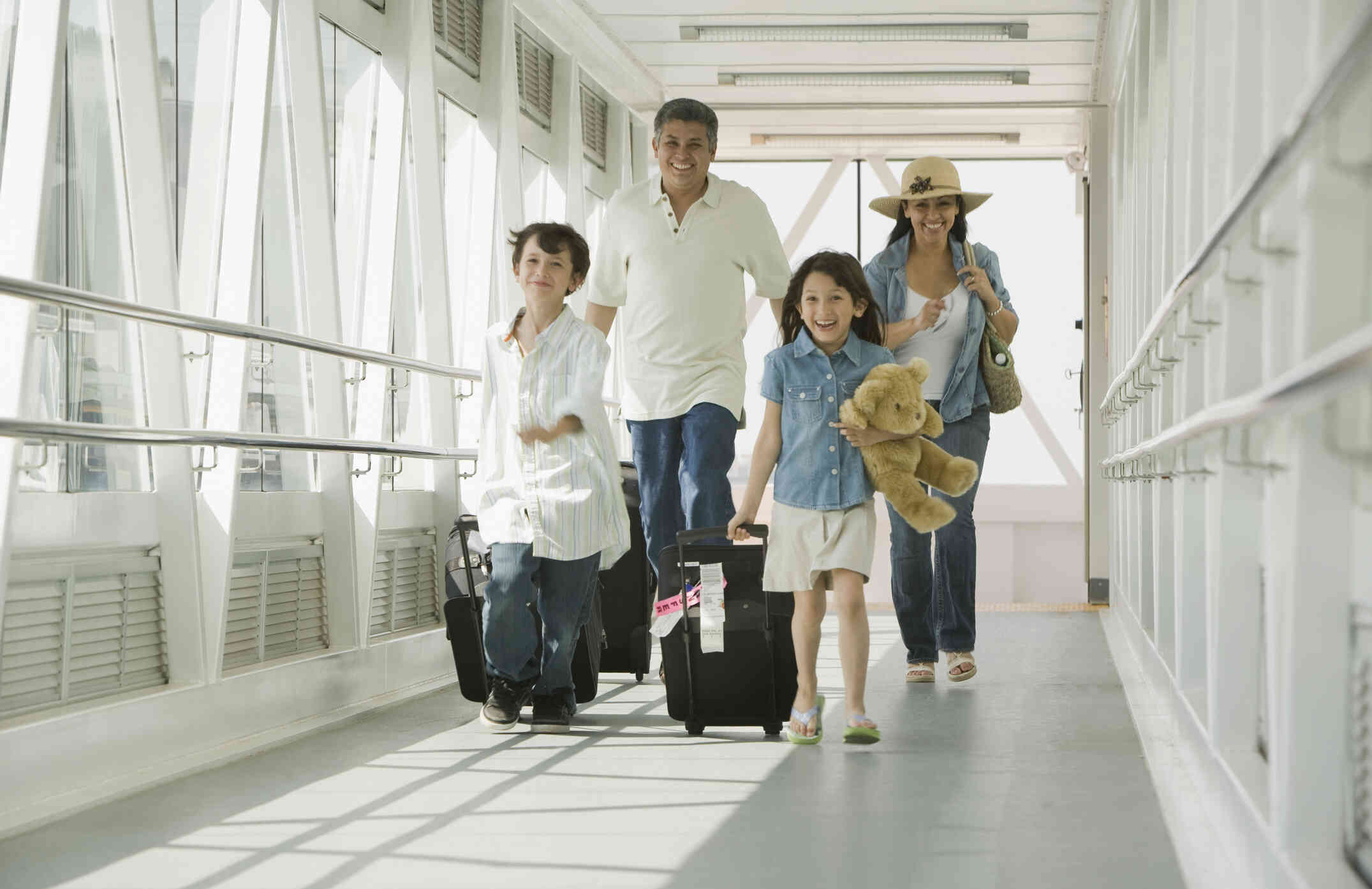 A man and a woman wearing a sunhat smile as they walk through an airport terminal. A young girl and boy walk in front of them smiling and pulling luggage.