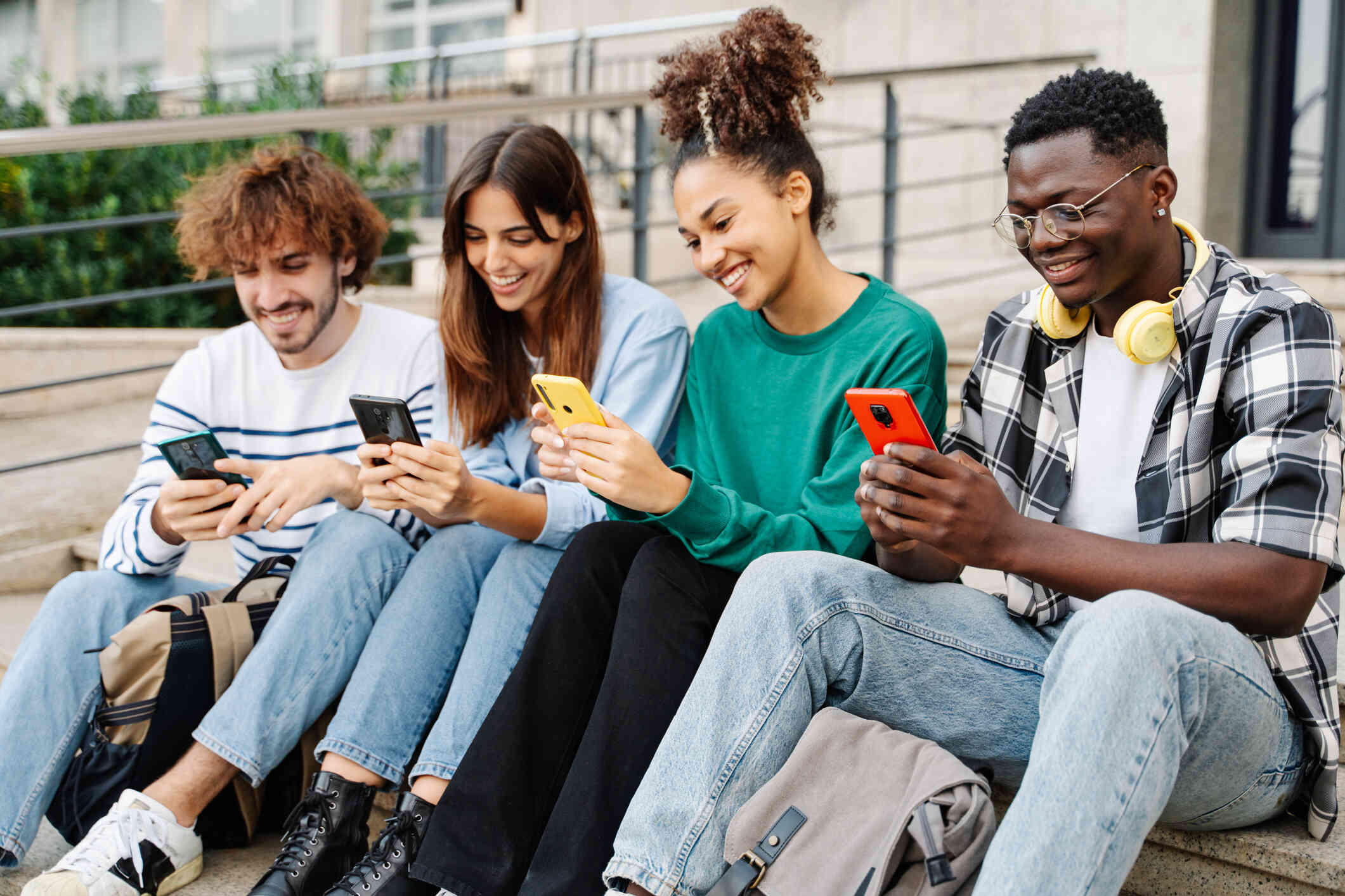 Four adult friends sit side by side outside while all smiling down at their phones in their hands.