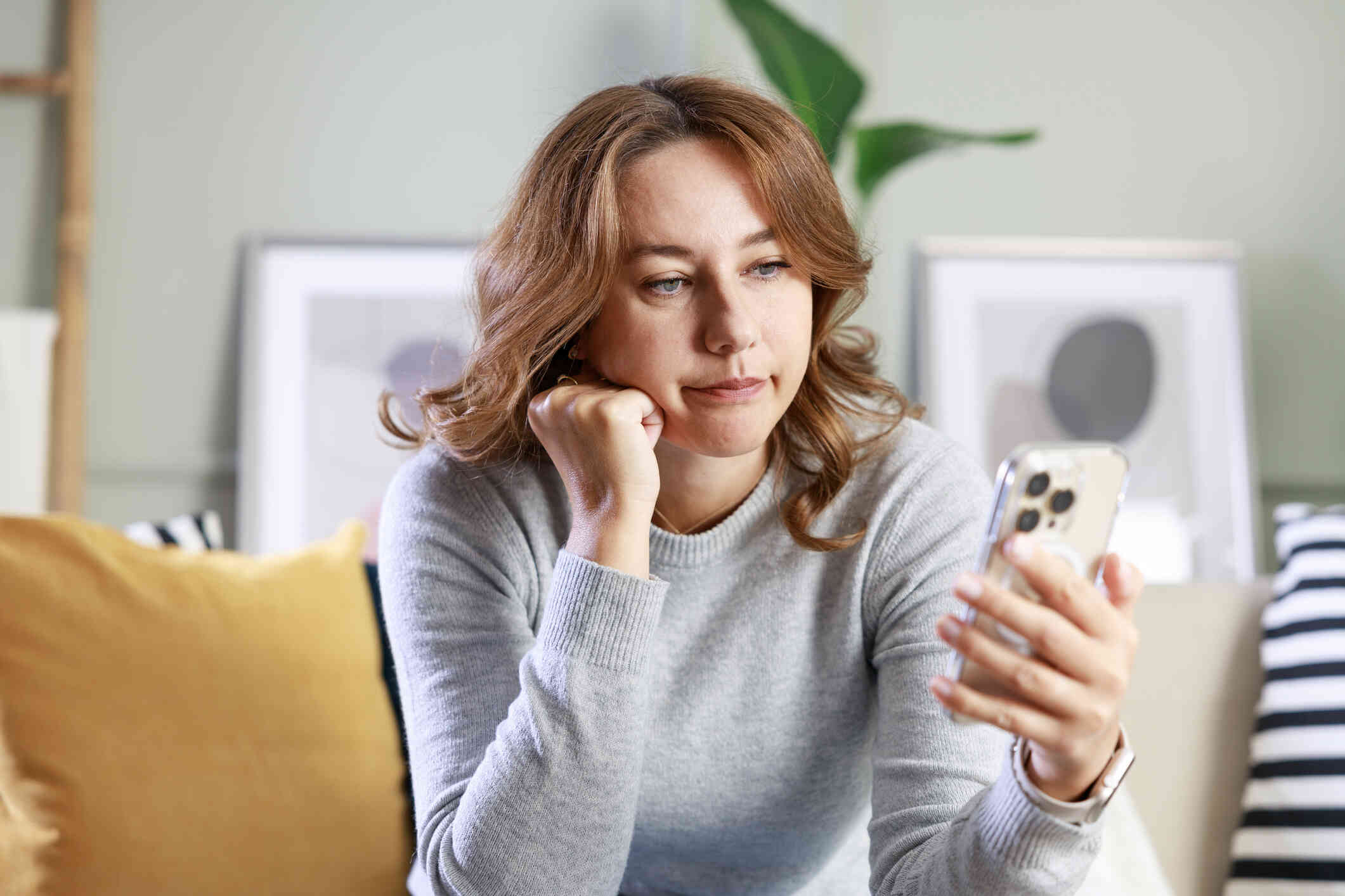 A woman looks upset as she sits on her couch and leans forward with her head resting against her hand as she looks at her phone.