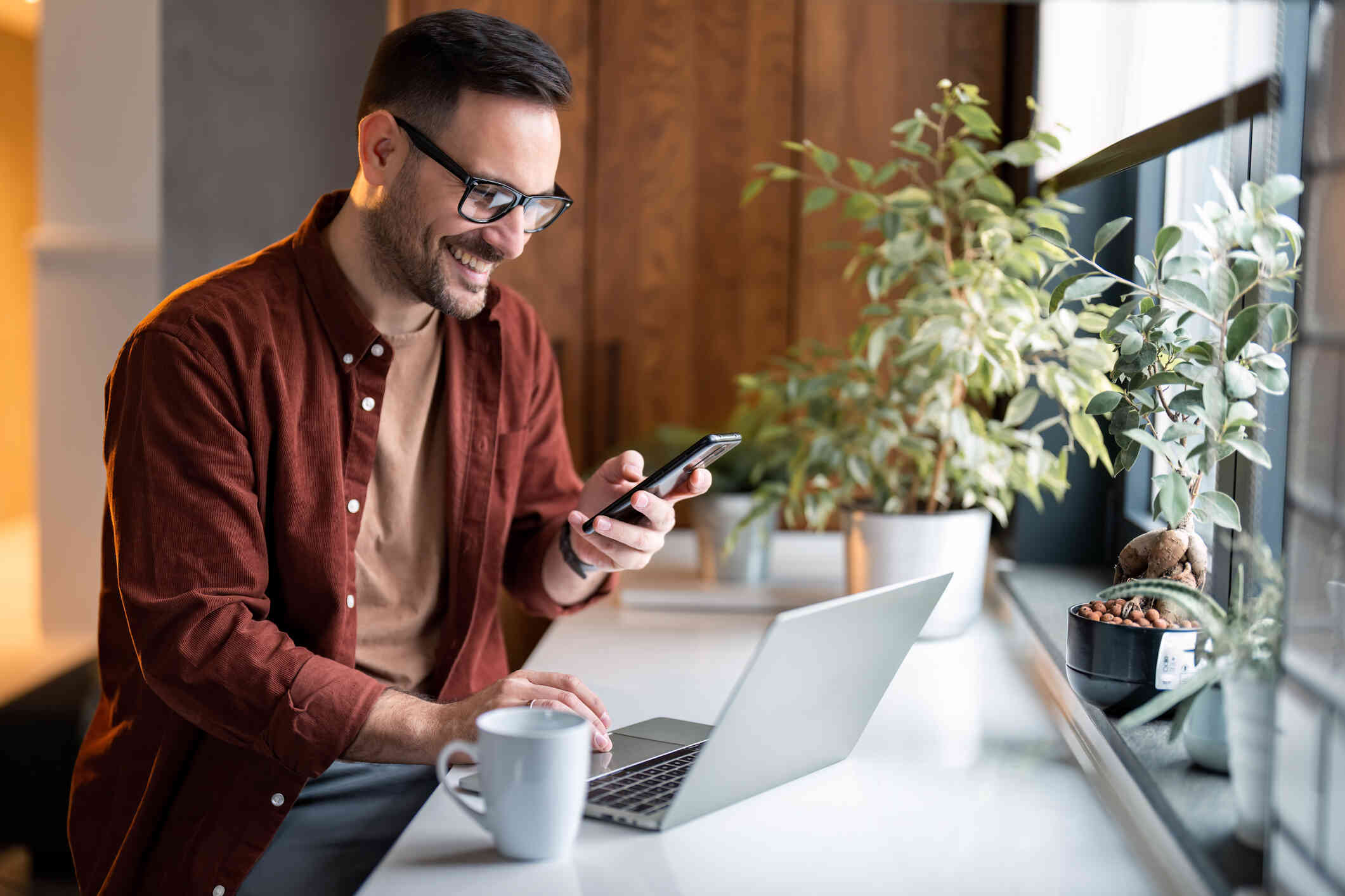 A man wearing glasses smiles as he holds a cell phone in his hand and sits at a counter by a window with plants, a coffee mug, and a laptop on it.