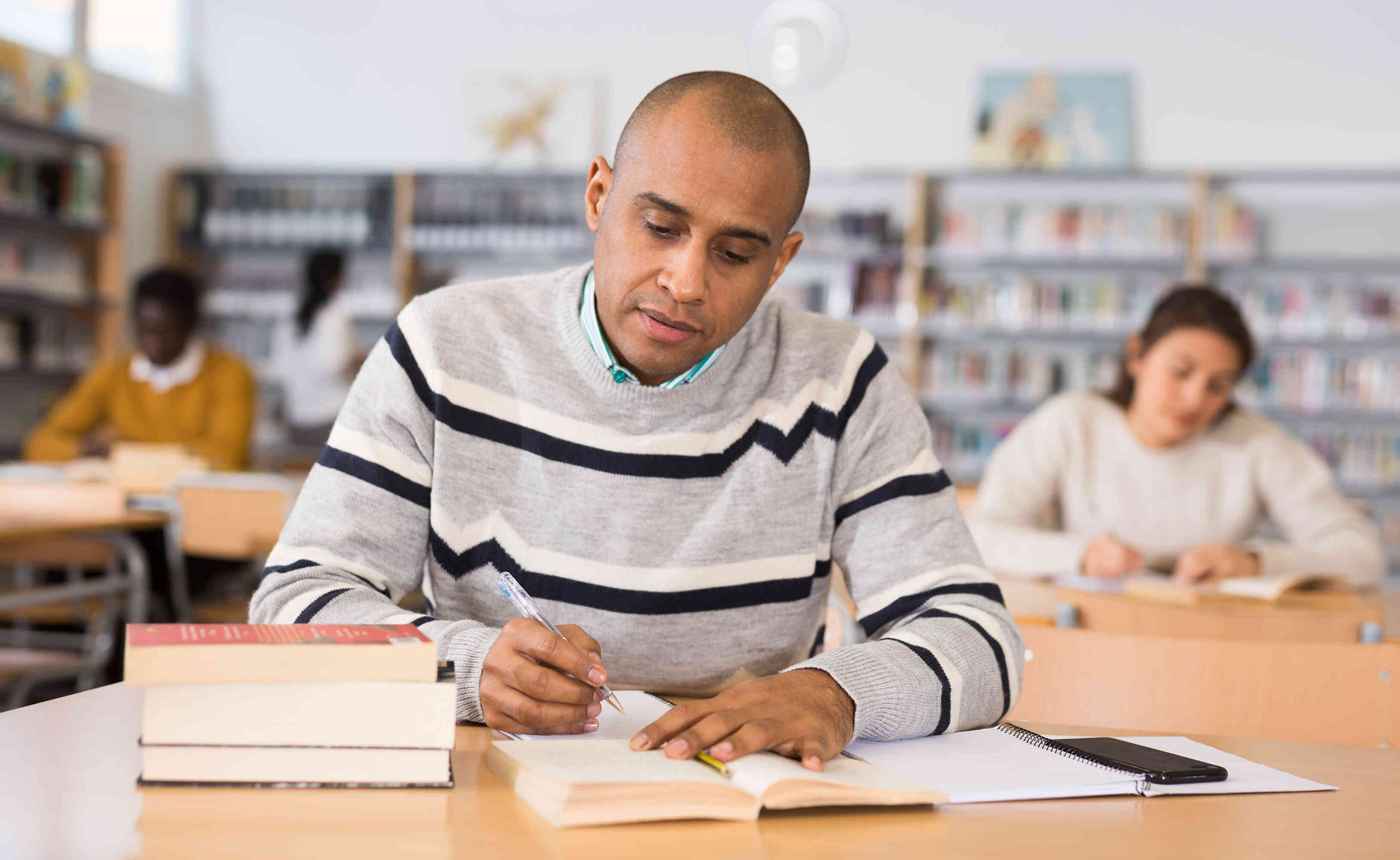 A man sits at a table in the library and looks through some books while taking notes.