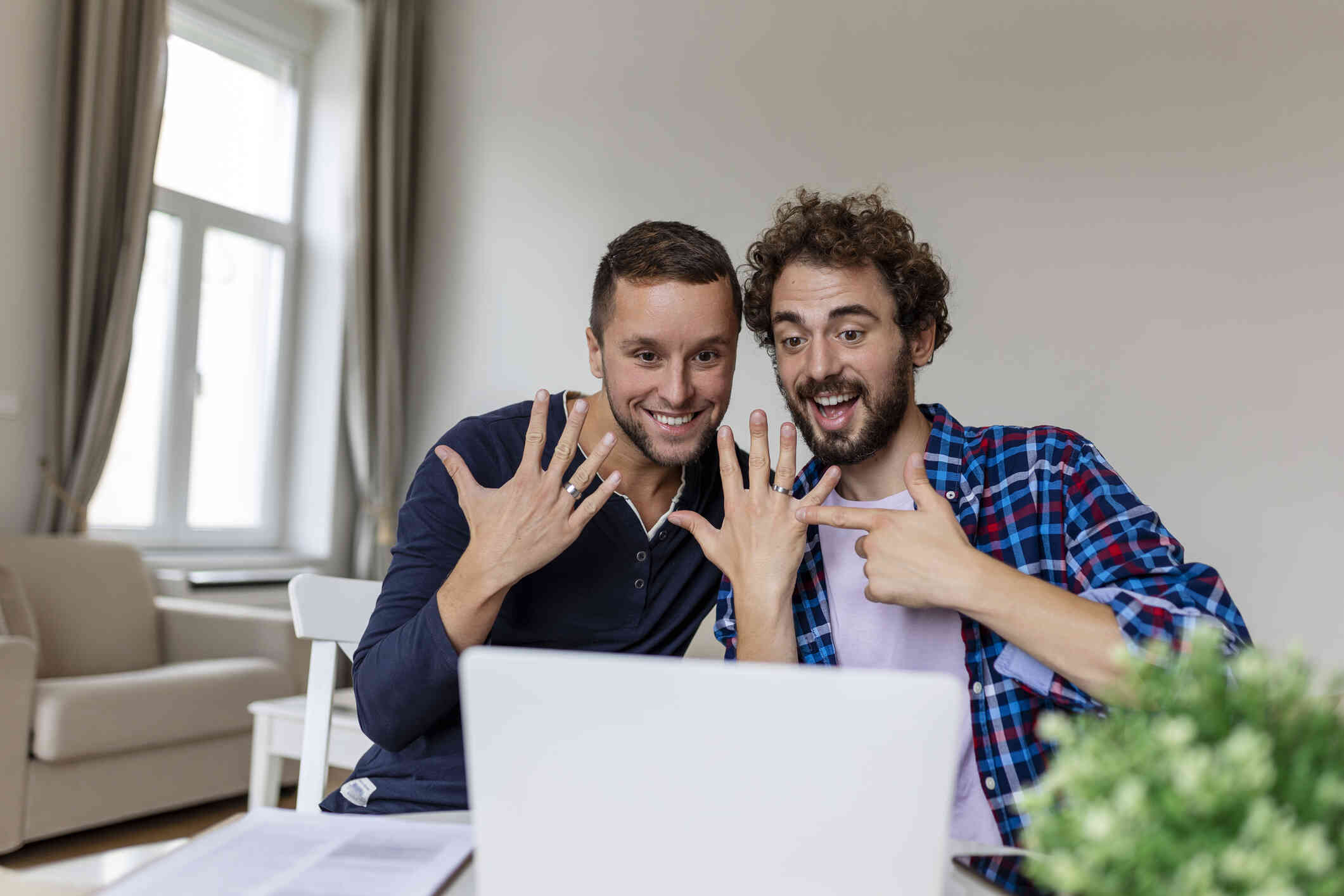 Two men sit next to each other in front of a laptop and smile as they hold up and point to their hands which have engagement rings on them.