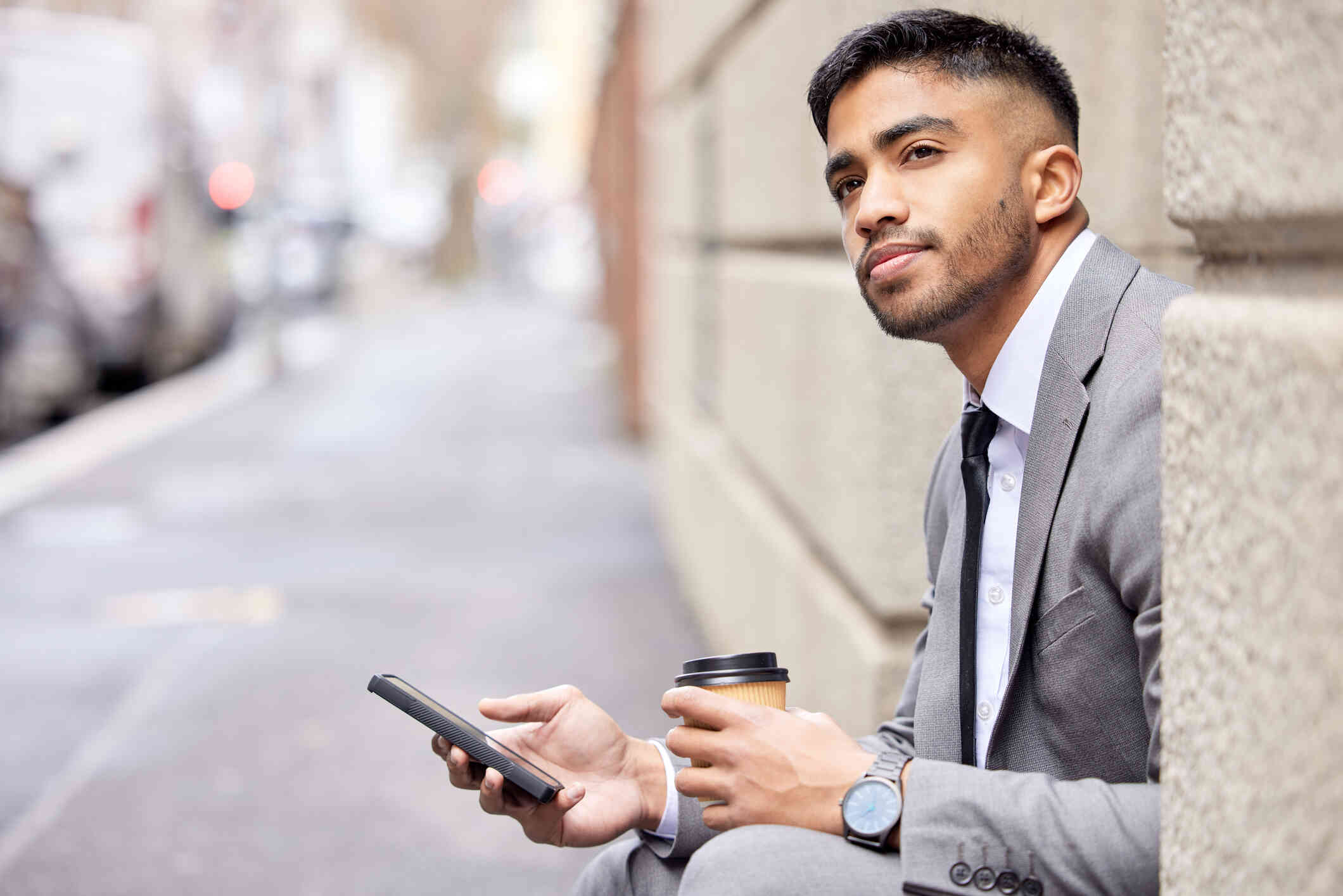 A man in a suit sits on a step outside and holds a cup of to-go coffee while holding a cellphone in the other hand and gazing off while deep in thought.