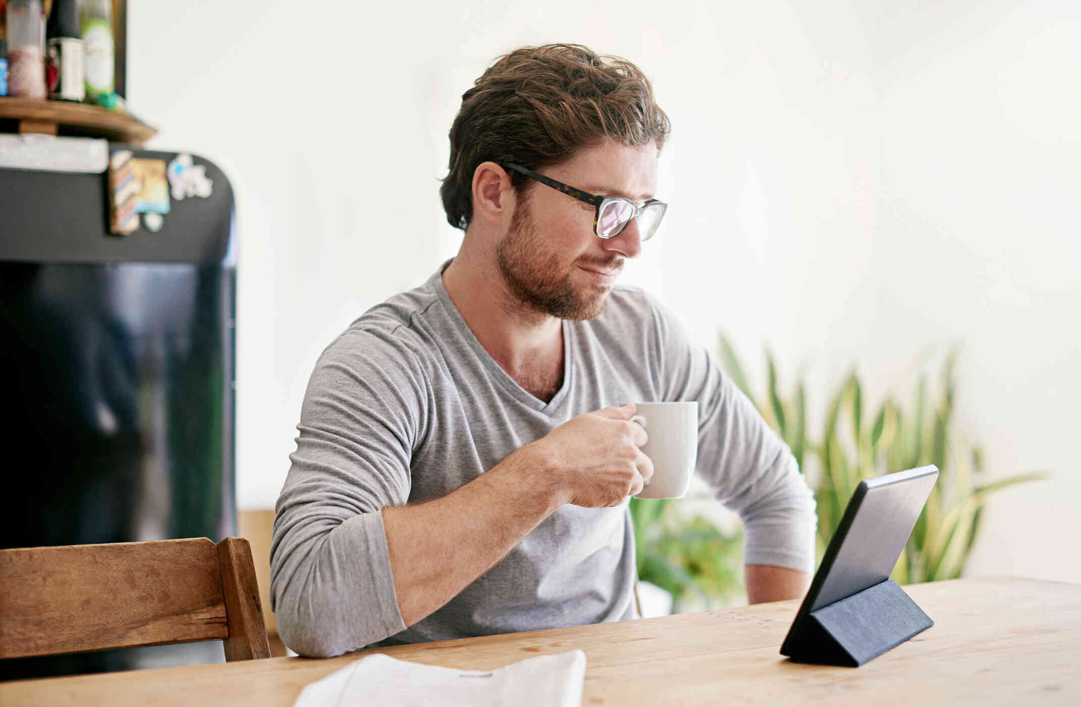 A man in a grey shirt sits at the kitchen table and holds a cup of coffee while smirking down at the tablet on the table infront of him.