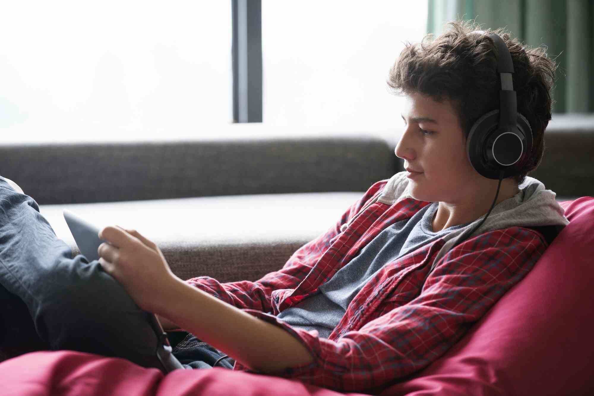 A teenage boy sits comfortably on a couch, wearing headphones while using his tablet.