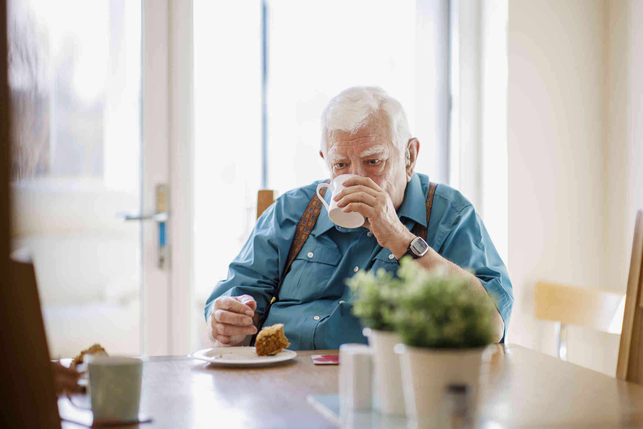 An elderly man in a blue button down shirt and suspenders sips from a white coffee mug while sitting at the breakfast table with a plate of half eaten food.