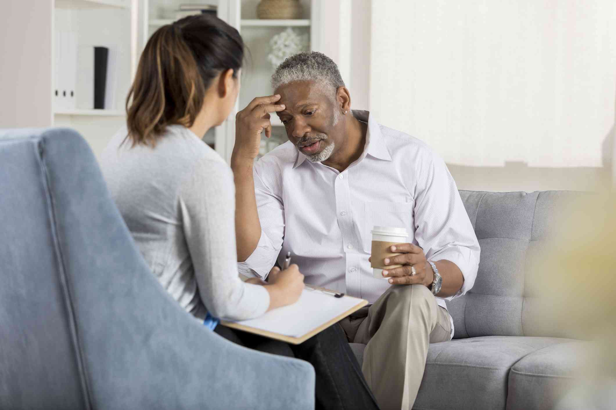 A middle aged man looks worried as he sits on a couch with a to-go cup of coffee in his hand and talks to the female therapist sitting across form him.