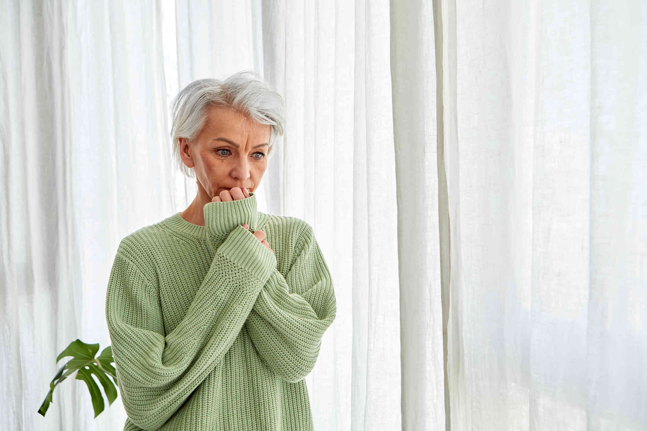 A mature woman in a green sweater looks stressed as she stands in her home and presses her hand to her face.