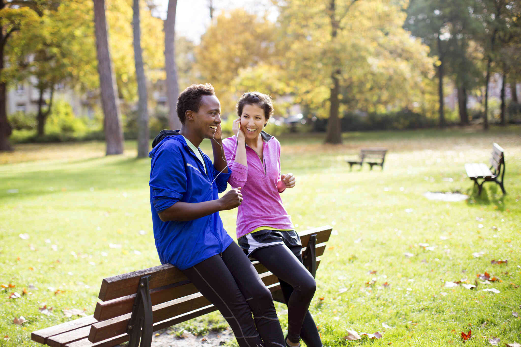 A male and female couple lean against a park bench on a sunny day and smile while shearing headphones.