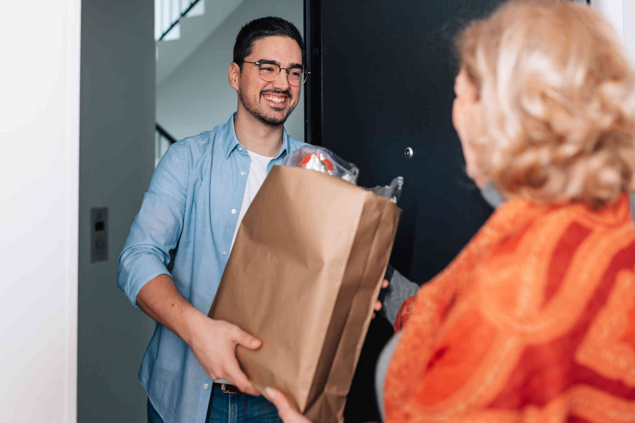 A man carries a paper bag of groceries through the front door with a smile as an older woman greets him.