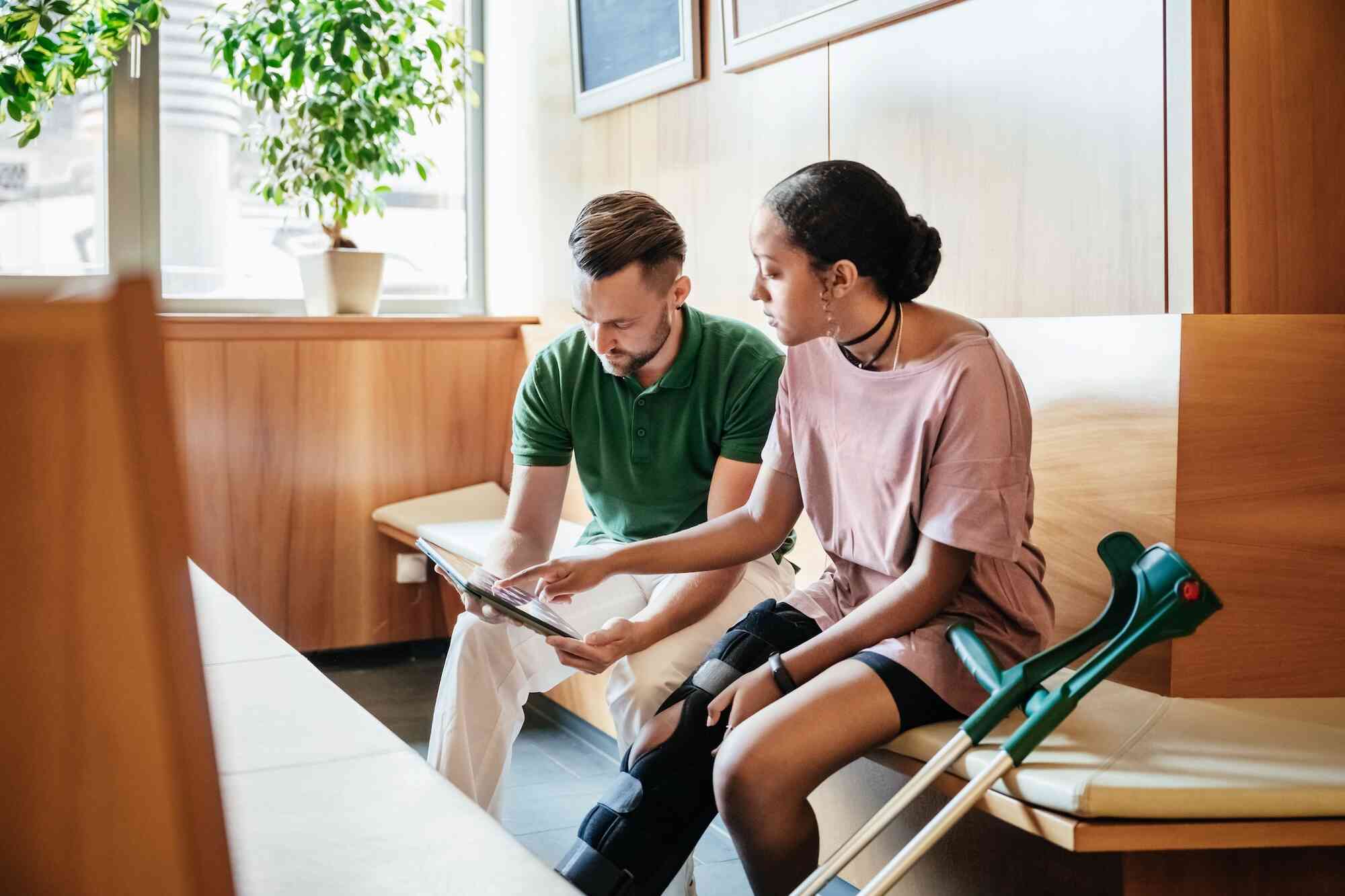 A teenage girl with a leg cast and her dad sit on a wooden chair, both checking the same tablet.