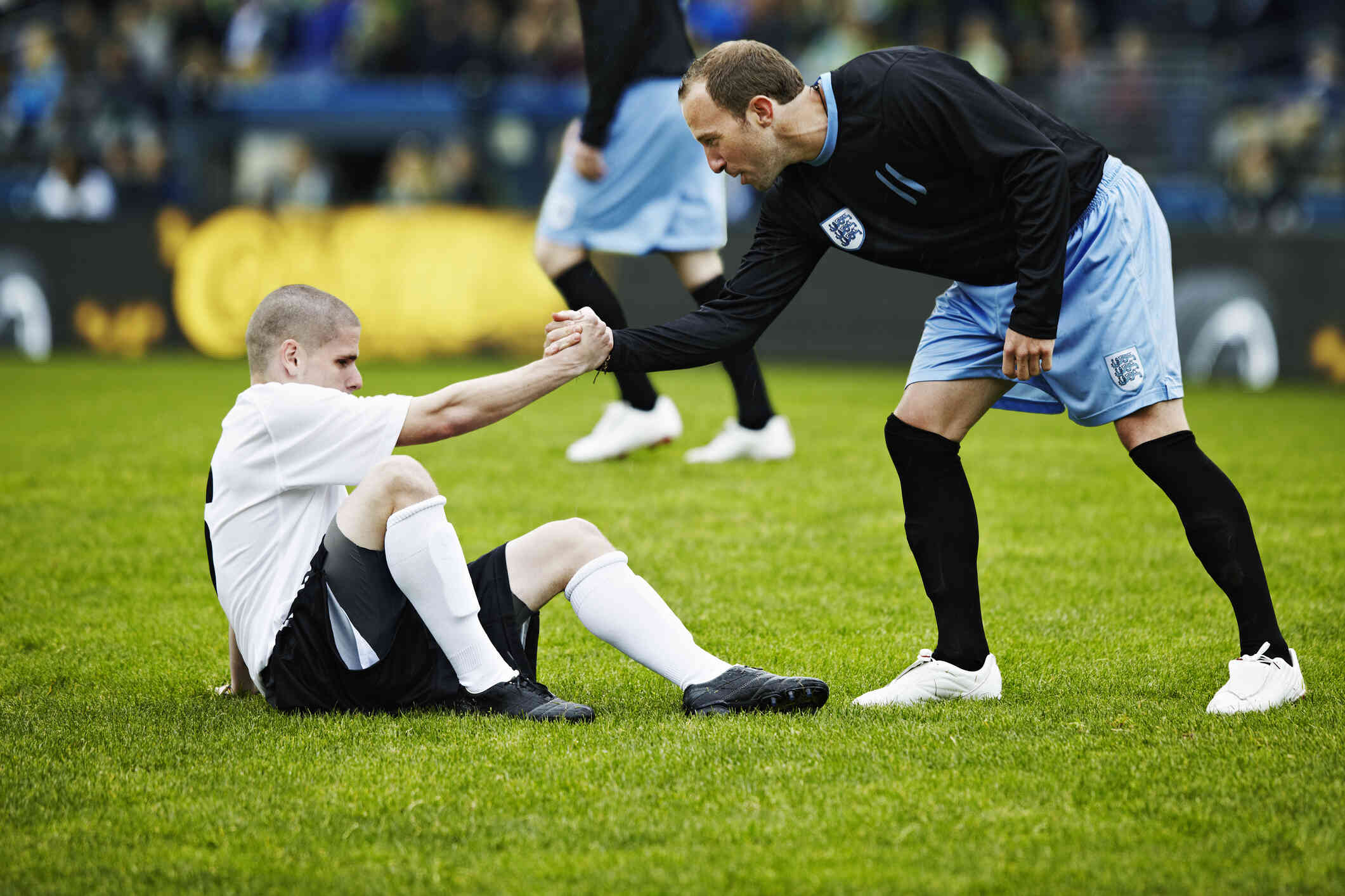 A male soccer player on a soccer field  leans down and holds the hand of a fallen player to help him up.