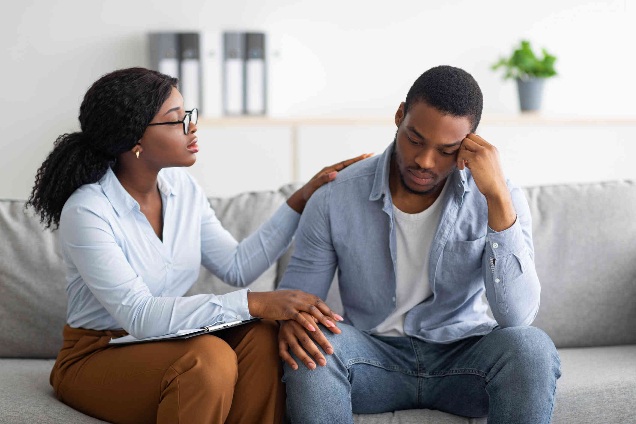 A man sits on the couch with his head resting sadly on his hand as his female partner sits next to him and tries to comfort him.