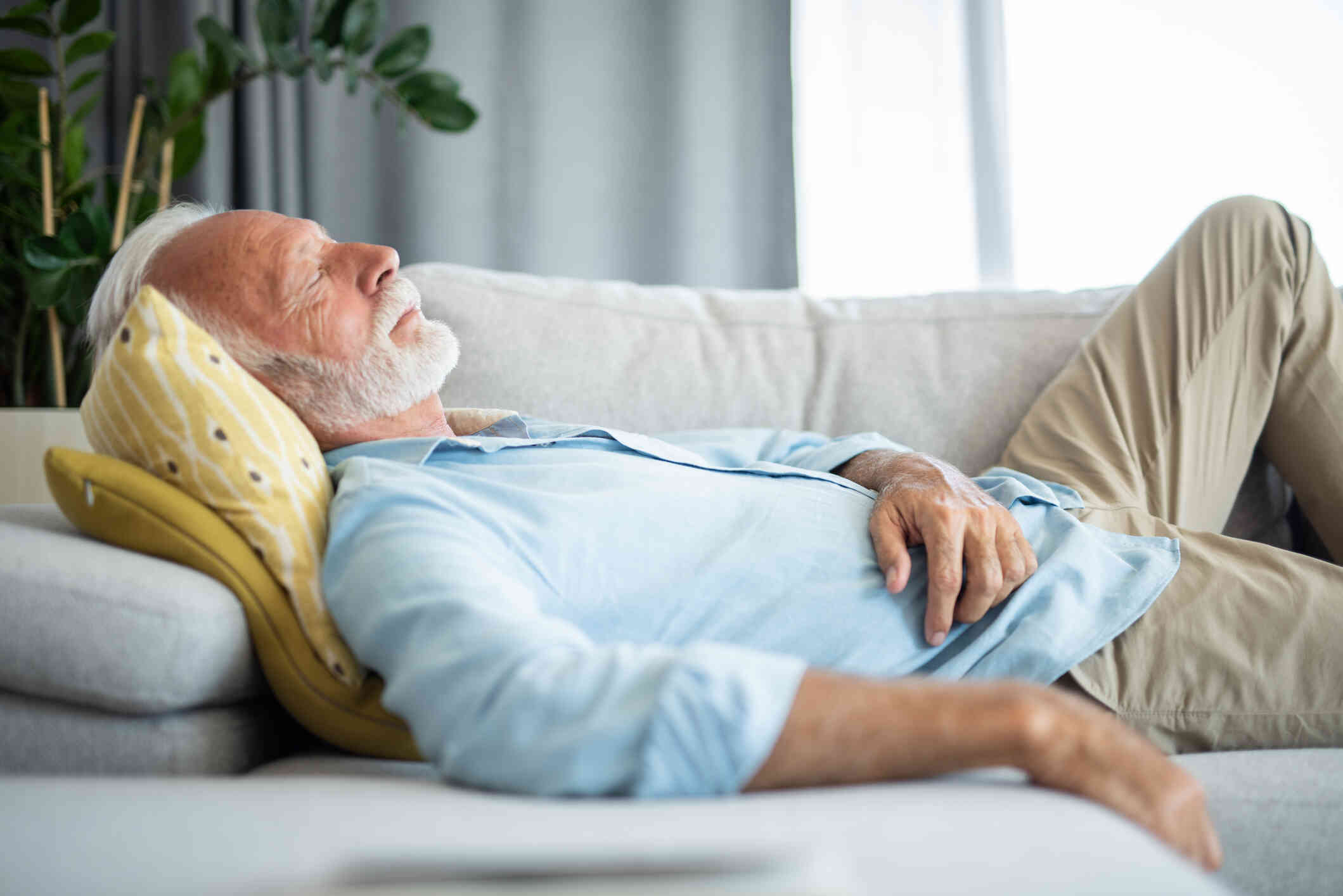 An older man in a blue shirt lies on a couch with his eyes closed while laying his hand on his stomach