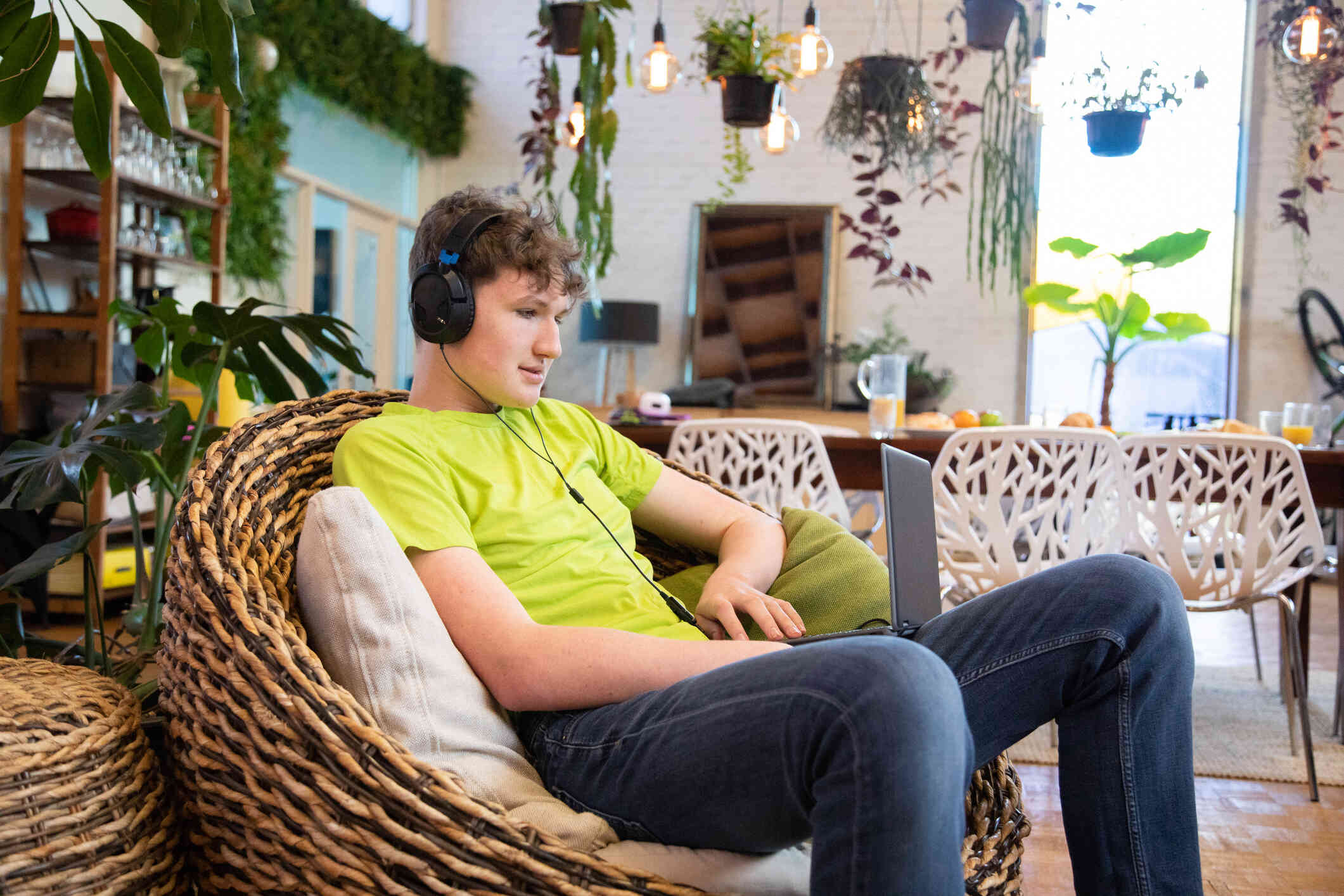 A teen boy in a green shirt sits in a wicker chair while wearing headphones and looks down at the laptop open in his lap.