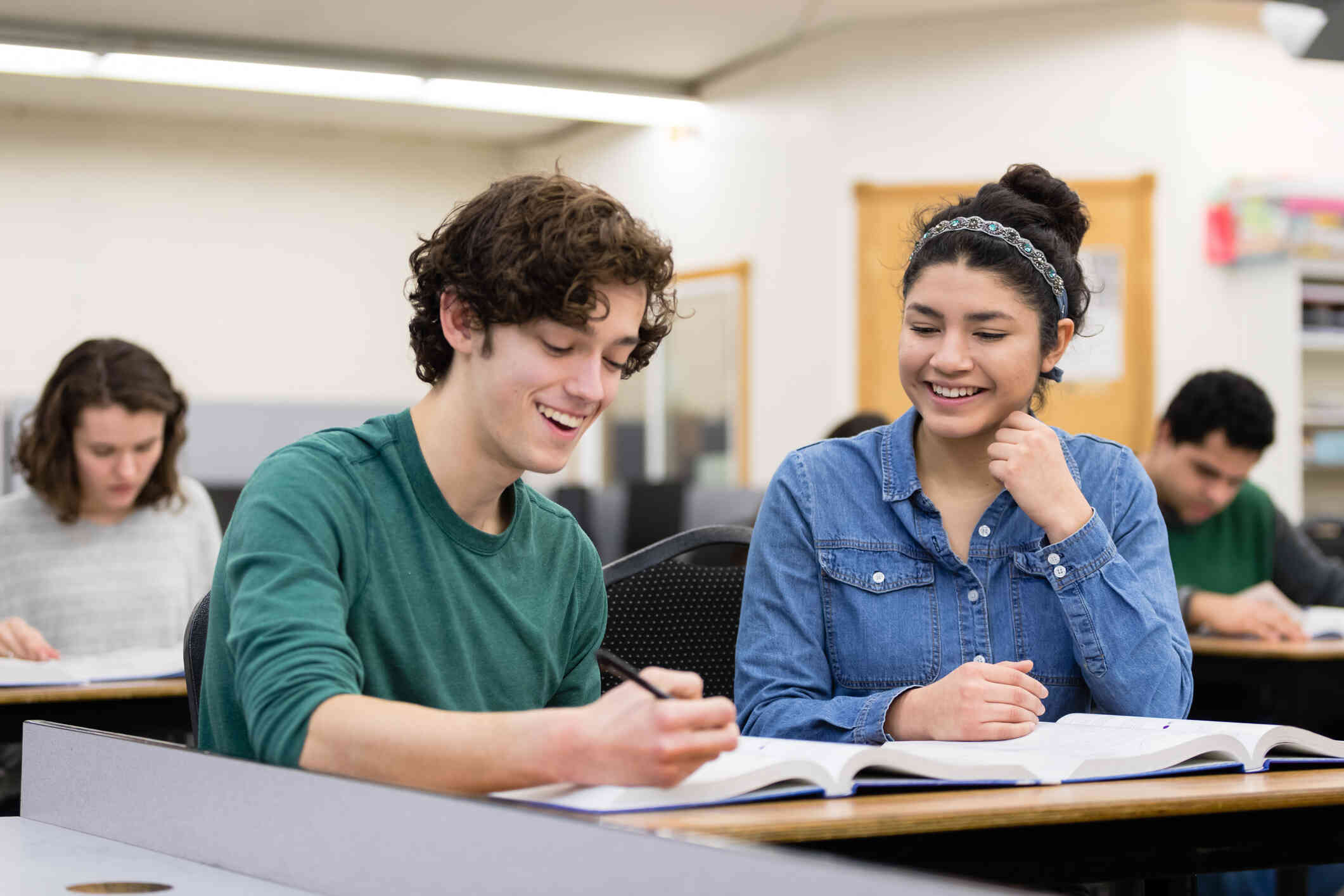 A young man in a green shirt and a young woman in a denim shirt sit next to each other at a desk smiling as they write in a book which is in front of them.