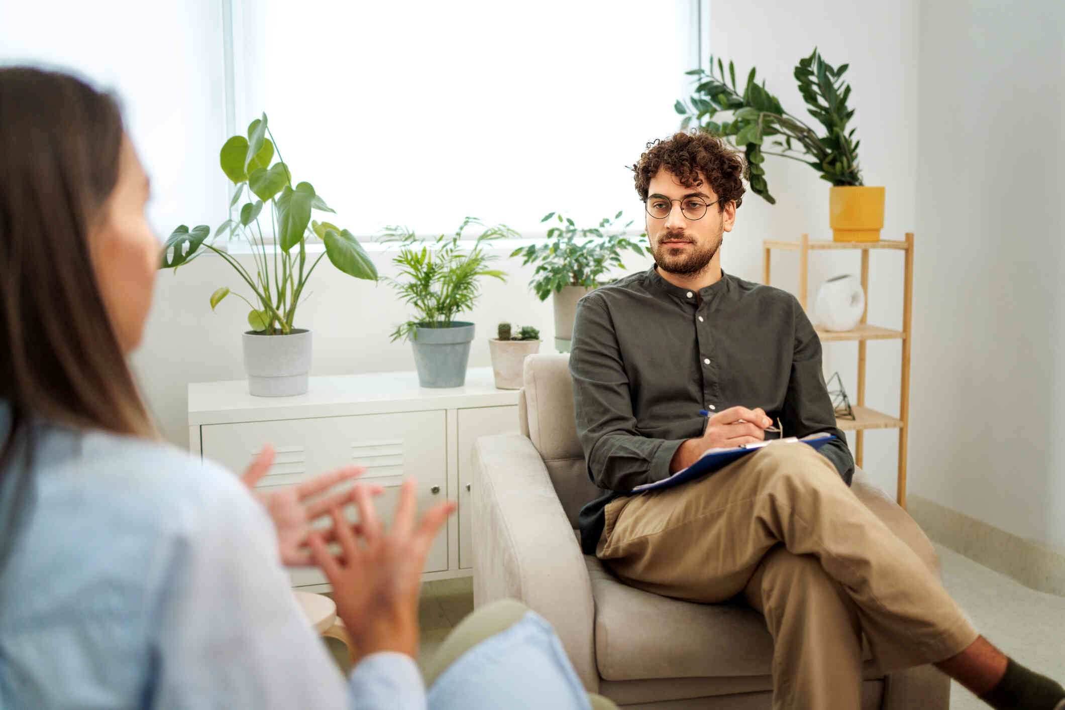 A male therapist in a button down shirt sits in a chair with a clipboard and pen and listens to his patient talk during a therapy session.