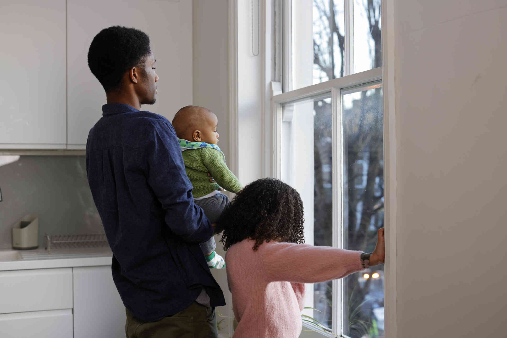 A dad holding his toddler stands next to his daughter as they gaze out of a window in their home.