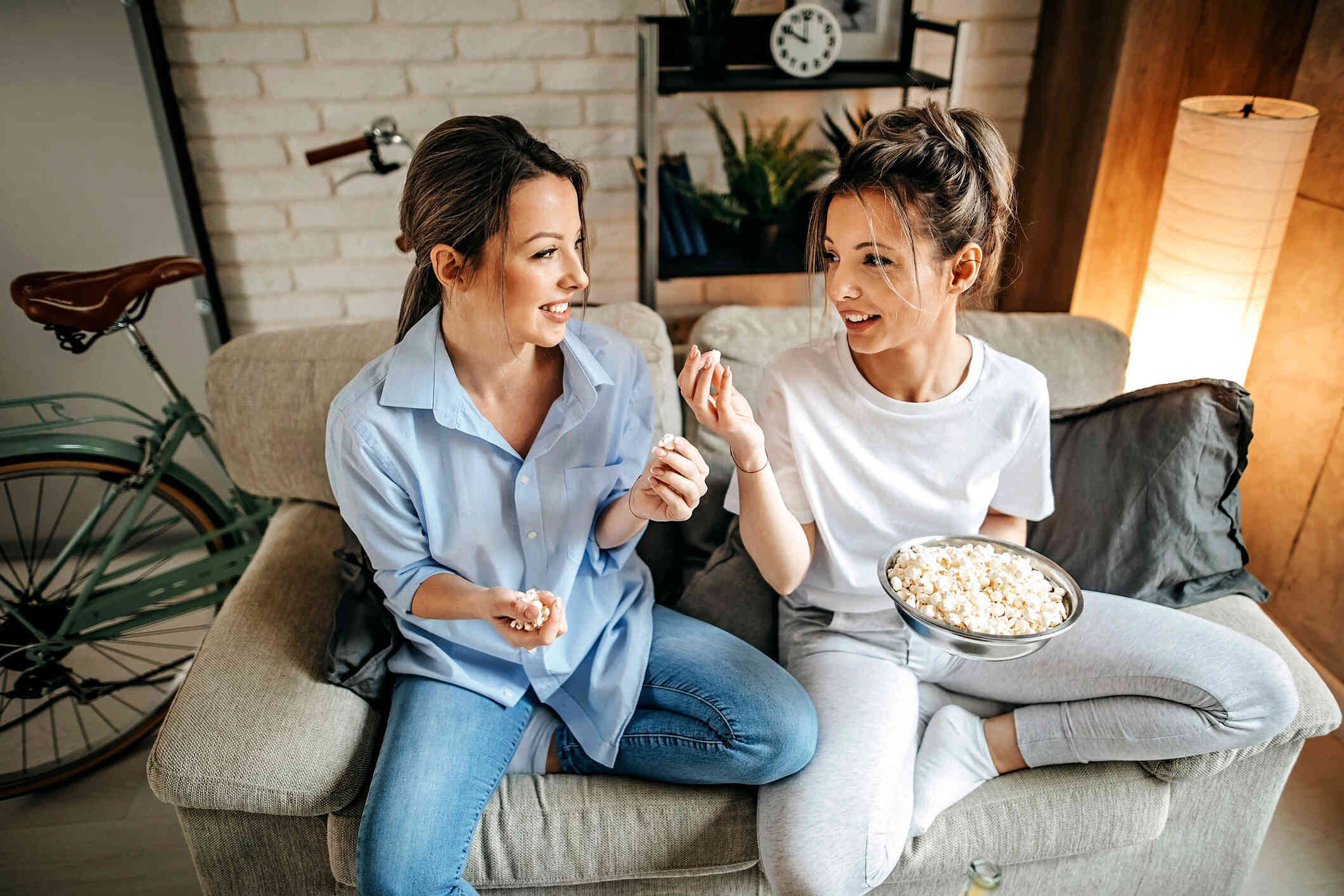 Two female adult friends sit next to each other on the couch while sharing a bowl of popcorn and smiling.