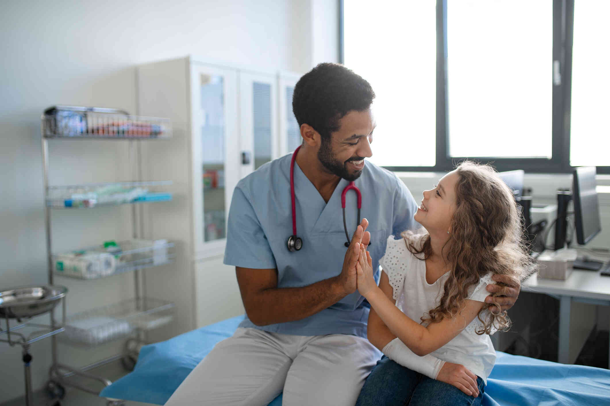 A man in scrubs sits next to a young girl in a doctors office and smiles as they high-five.