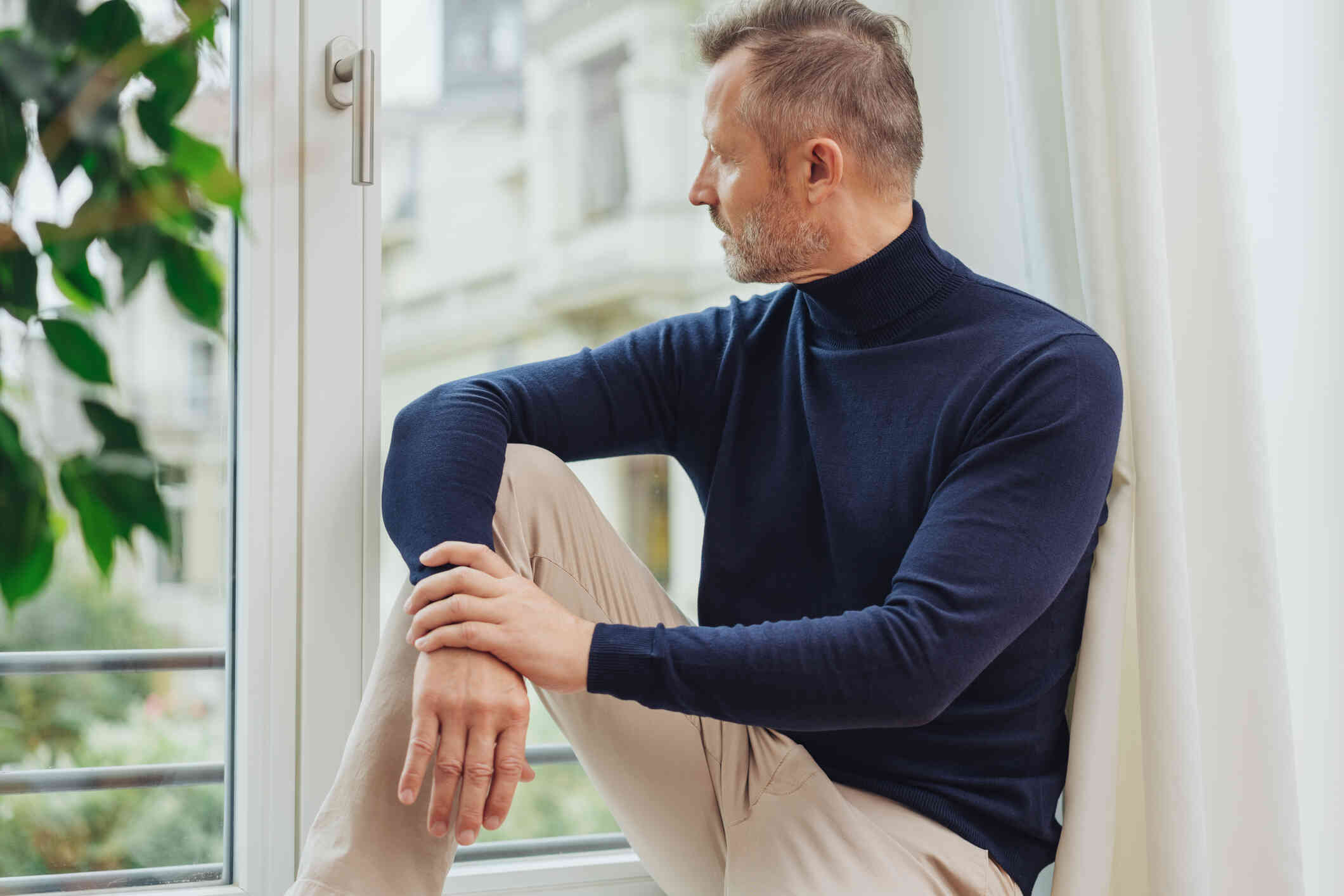 A man in a blue turtleneck sits in a wondow sill and gazes out while deep in thought.