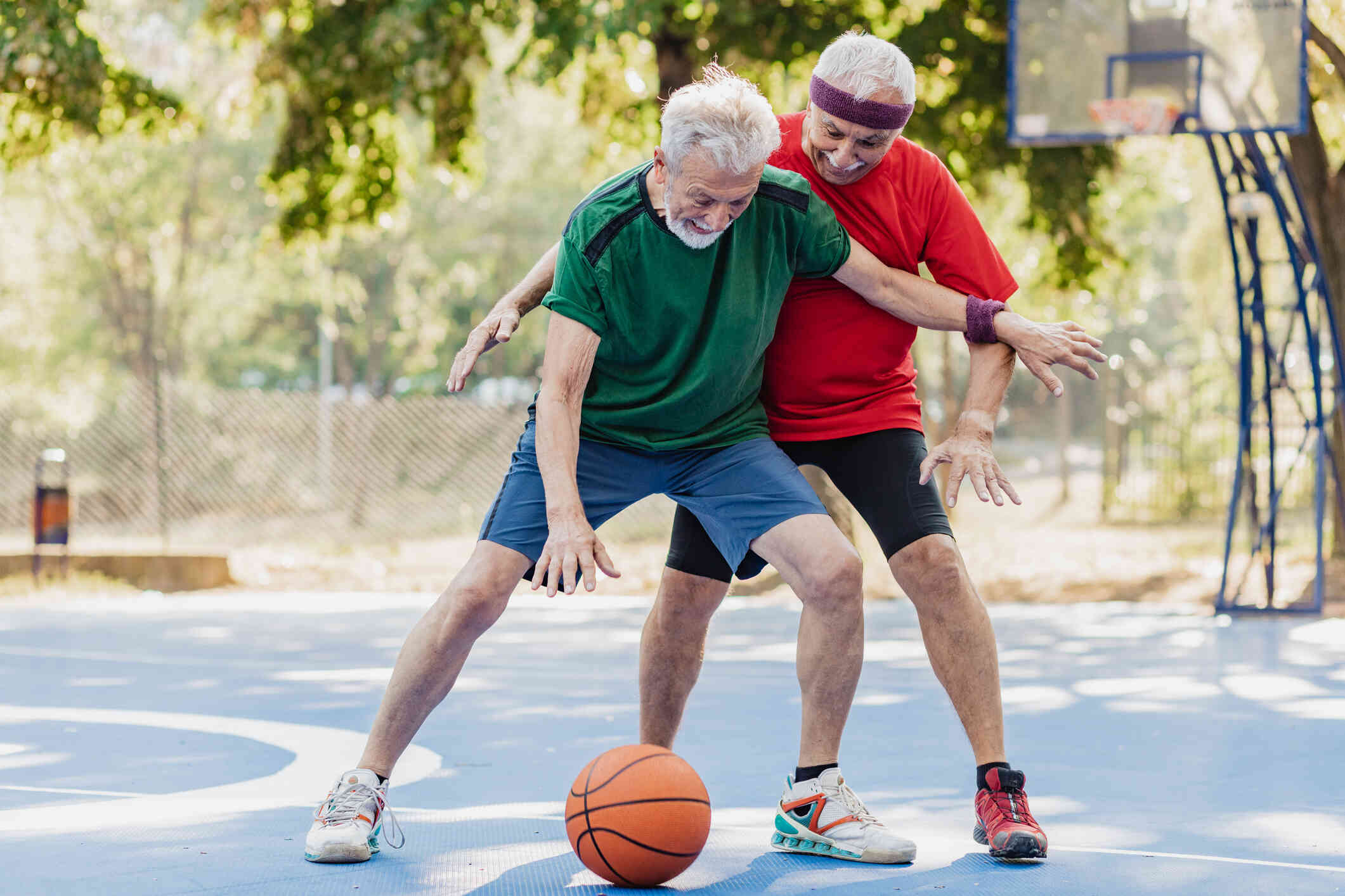 Two older men play basketball together on an outdoor court on a sunny day.