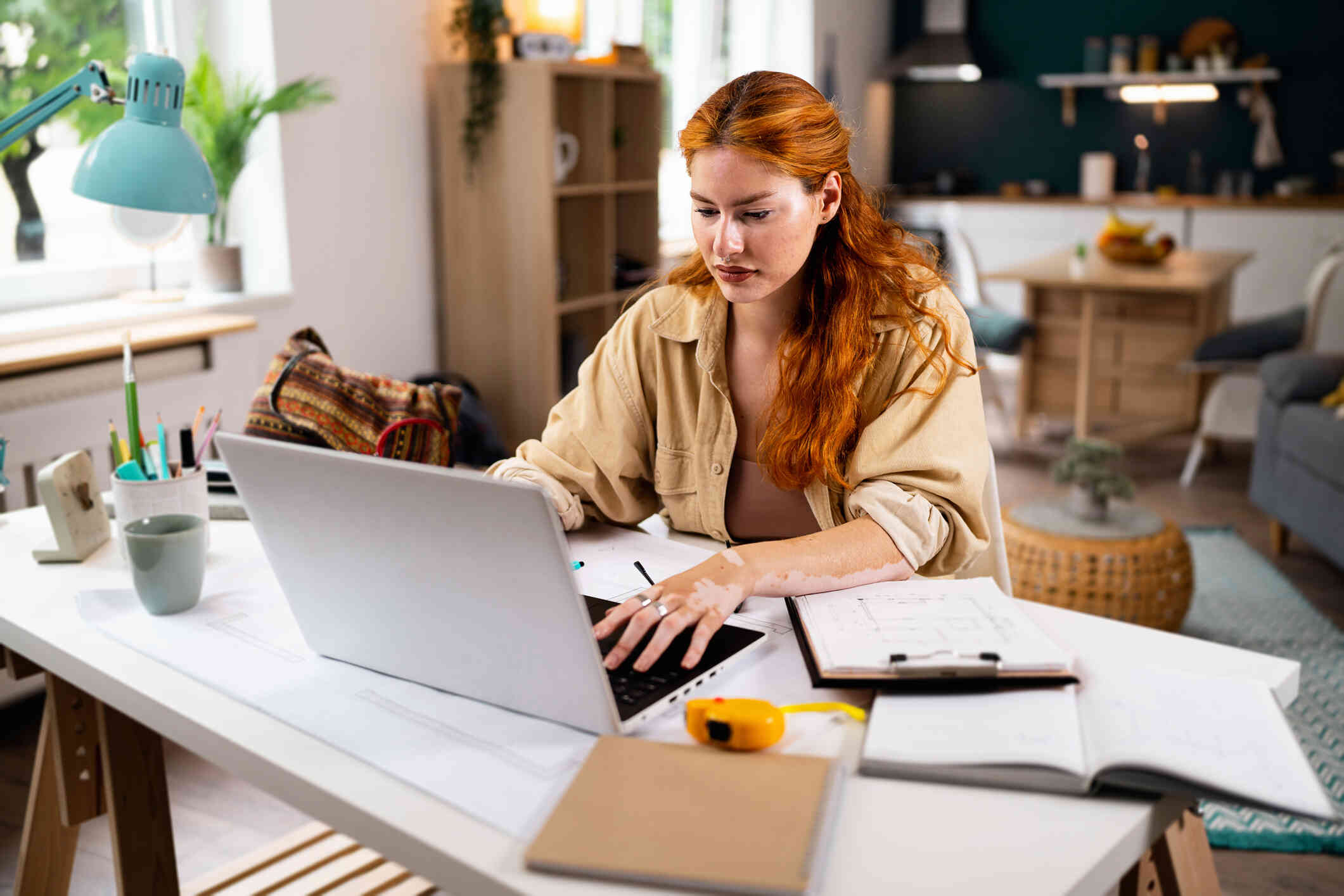 A woman in a tan shirt sits at her desk in her home and types on the laptop open on the table infront of her with a serious expression.