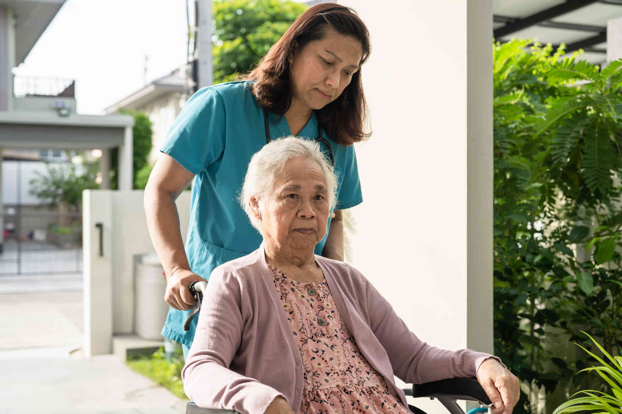 A nurse in green scrubs pushes an elderly female dementia patient in a wheelchair outside on a sunny day.